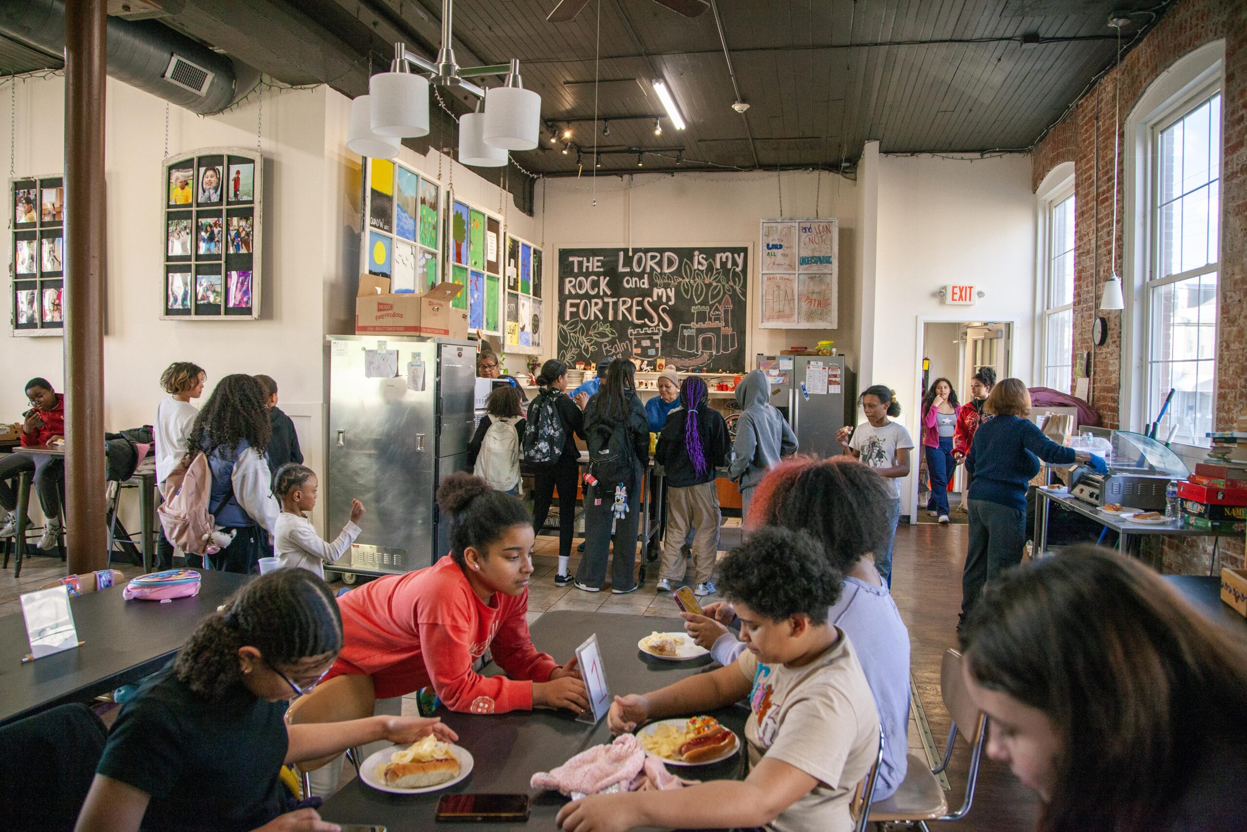 A lively community kitchen filled with children interacting, eating, and enjoying activities. In the background, colorful artwork adorns the walls, and a chalkboard displays an inspirational message. The space is bright and welcoming, showcasing a vibrant atmosphere of collaboration and engagement.