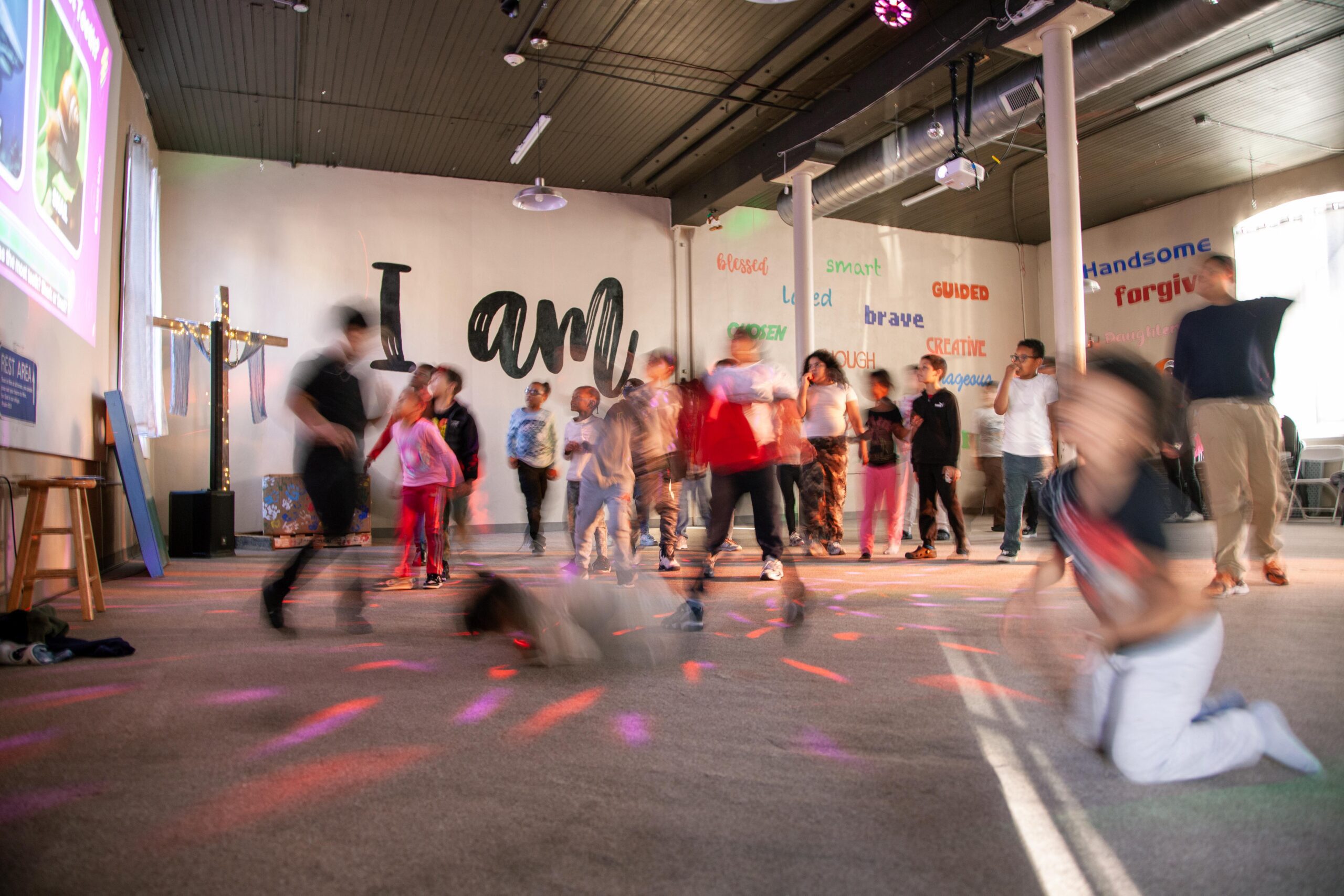 Children dancing in a lively indoor space with colorful lights, featuring a wall with motivational words and phrases.