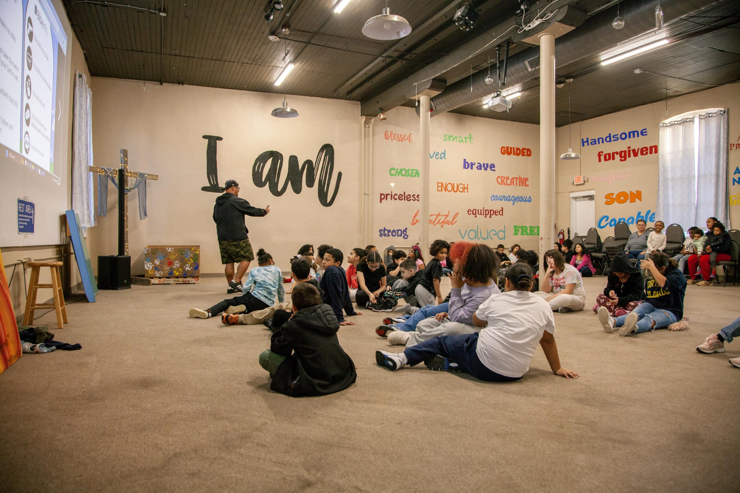 Children engaged in a group activity in a community center, with a speaker presenting in front of a large screen. The walls are decorated with positive affirmations like "I am," "brave," and "valued," creating an encouraging environment for youth development.