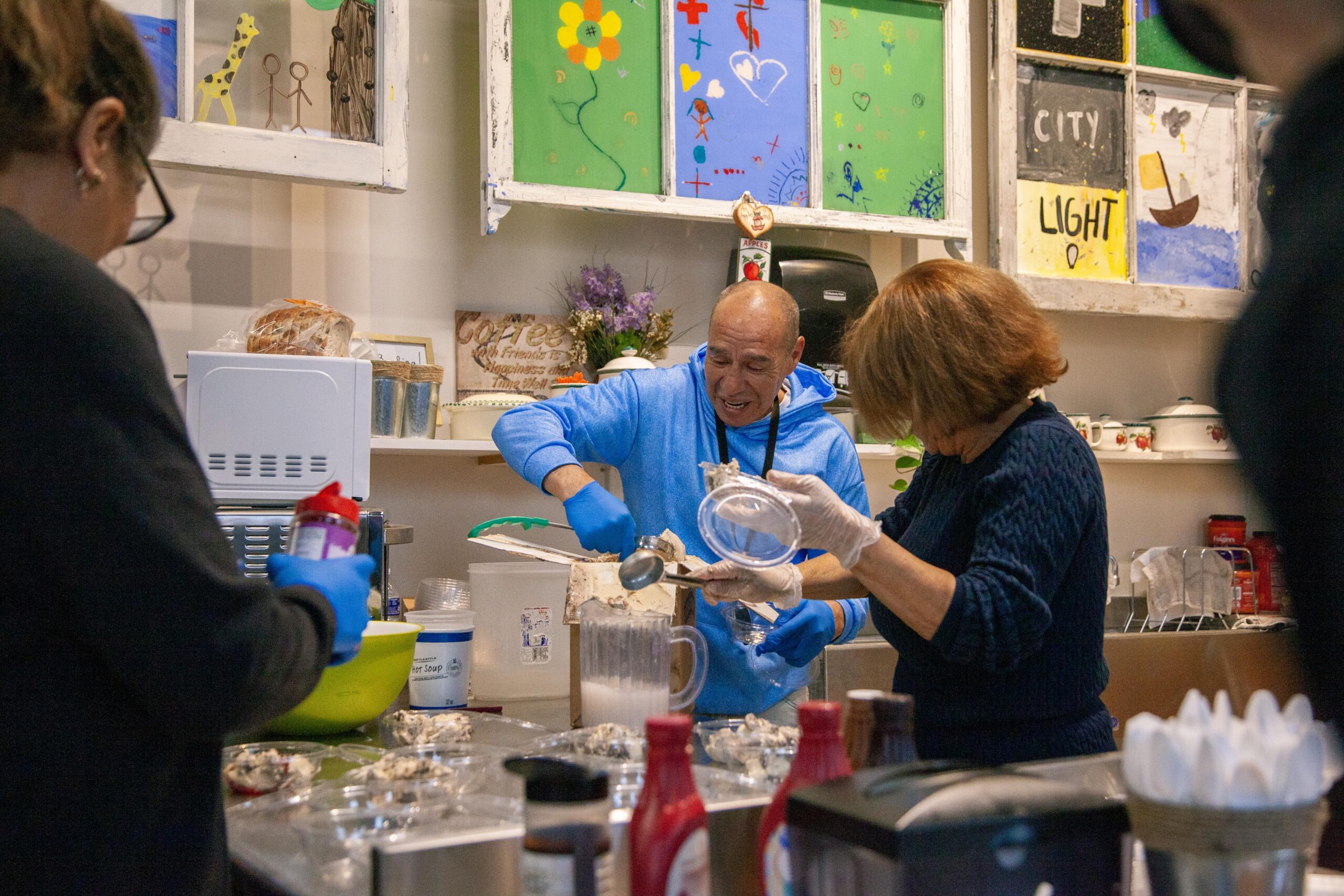 Two people preparing food in a kitchen setting, with one man in a blue hoodie scooping ingredients and a woman in a dark sweater holding a container. The kitchen features colorful artwork on the walls and various cooking supplies on the counter.