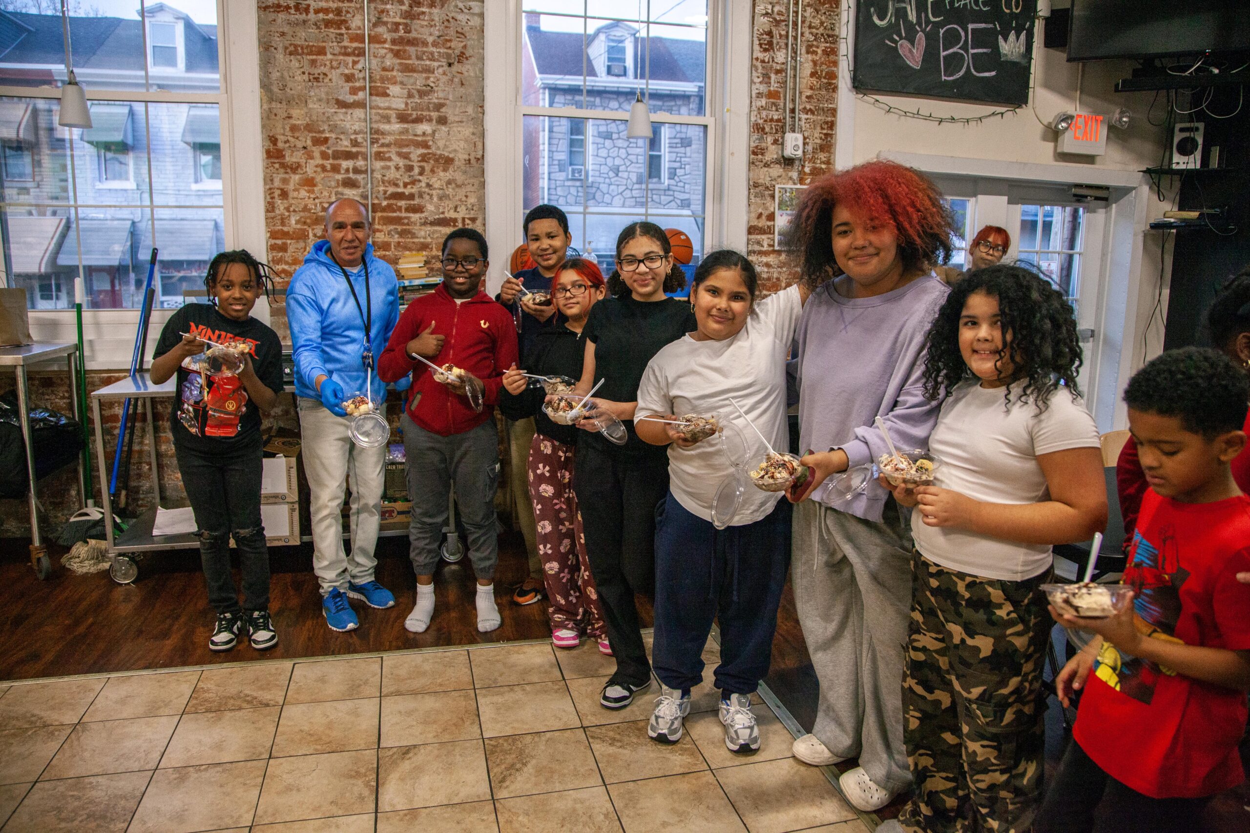 Group of children and adults smiling and holding dessert cups in a community center with a rustic brick backdrop, promoting teamwork and enjoyment in a fun, engaging environment.