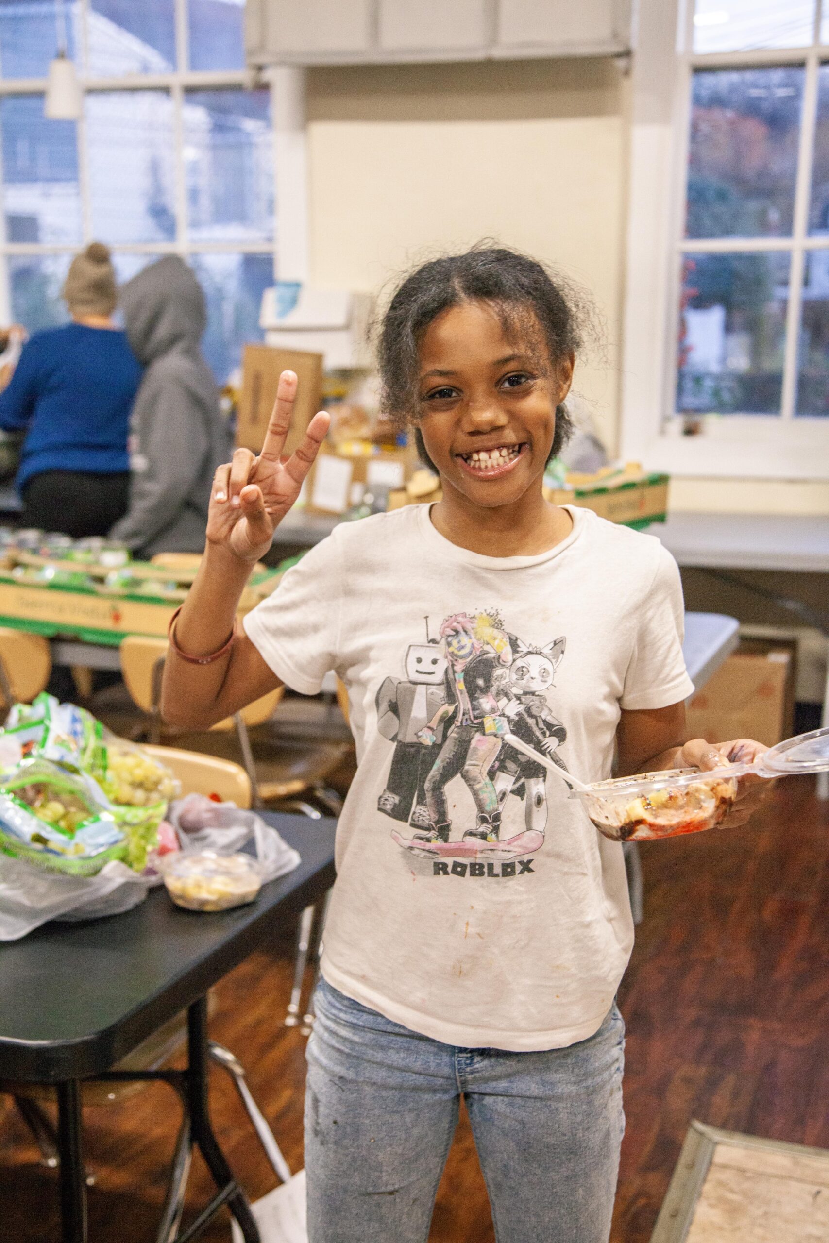 Smiling girl giving a peace sign while holding a food container, wearing a Roblox-themed t-shirt, in a community kitchen setting with food supplies in the background.