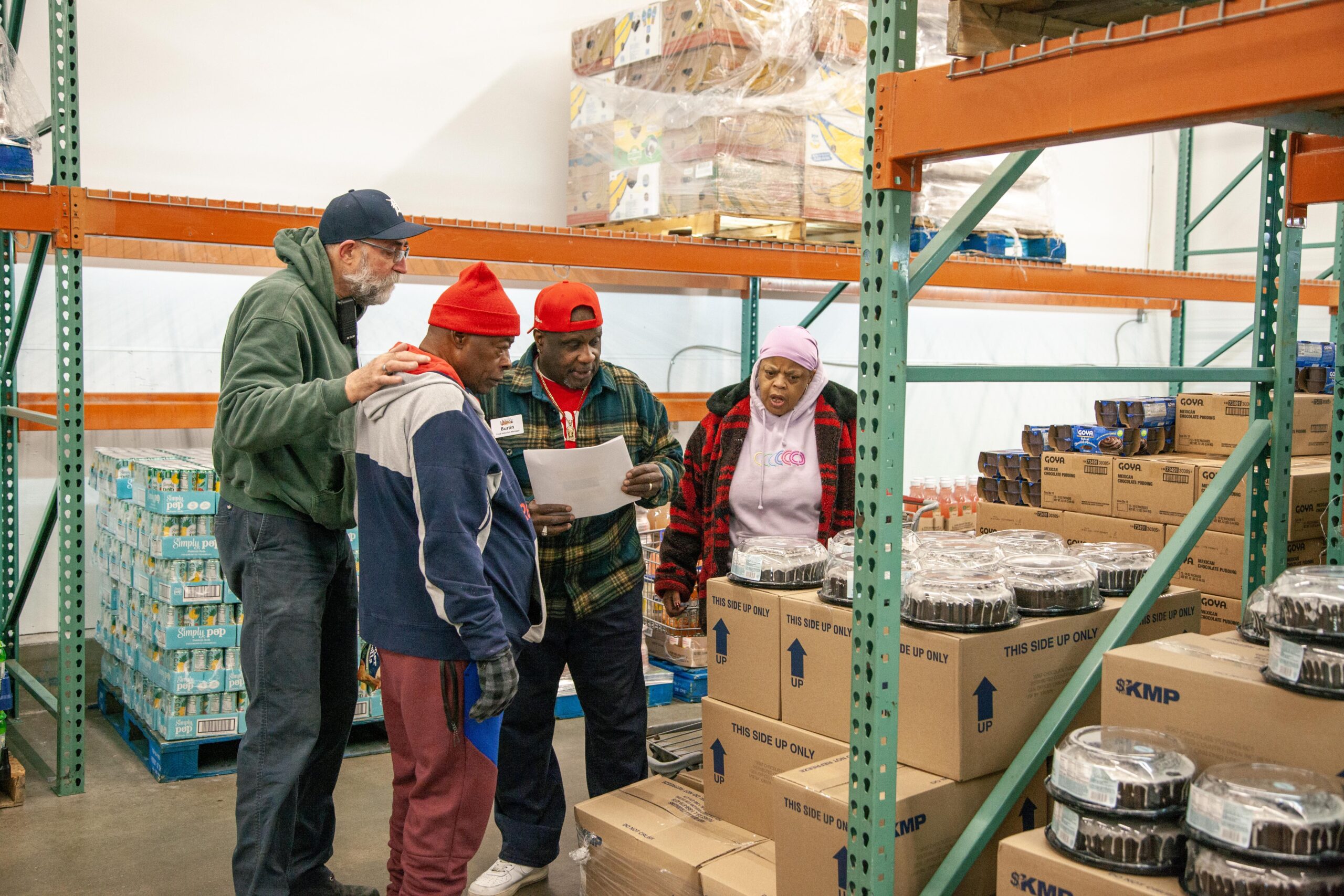 Group of four individuals collaborating in a food distribution warehouse, reviewing documents and discussing inventory. Shelves are stocked with boxes of food and beverages, highlighting the community effort in food assistance.