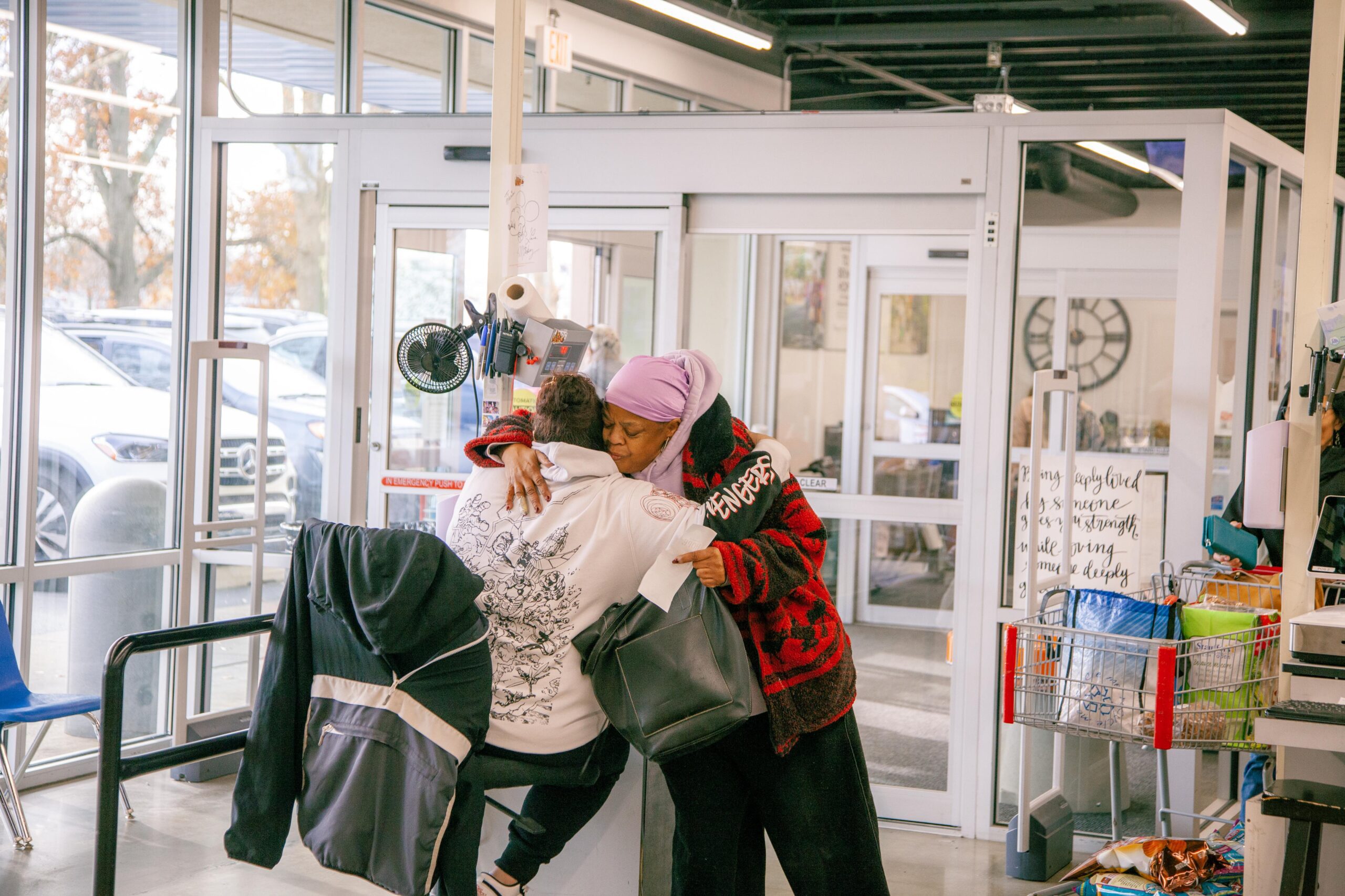 A heartwarming scene inside a store showing two women embracing, conveying a sense of connection and support. One woman is seated while the other stands beside her, both dressed in casual clothing. The store features large windows and a busy atmosphere with shopping carts visible in the background.