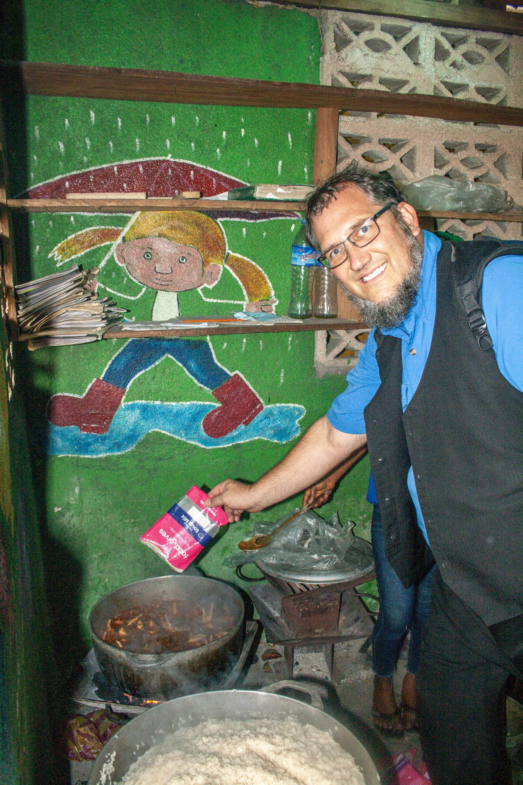 Man adding seasoning to a pot while cooking in a vibrant kitchen with a colorful mural of a child on the wall. The scene captures the essence of home-cooked meals and cultural traditions.