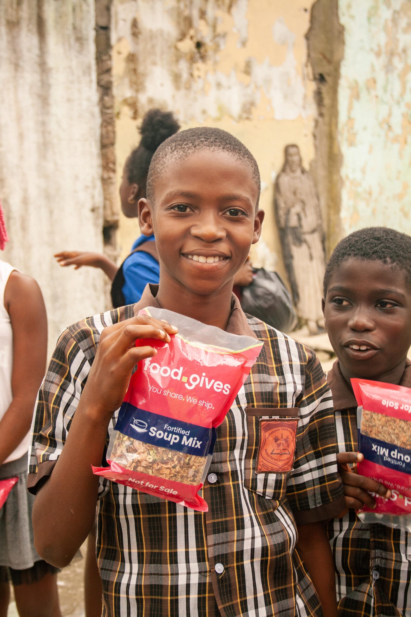 Smiling children holding packets of fortified soup mix from Food Gives, in a community setting with a weathered wall in the background.