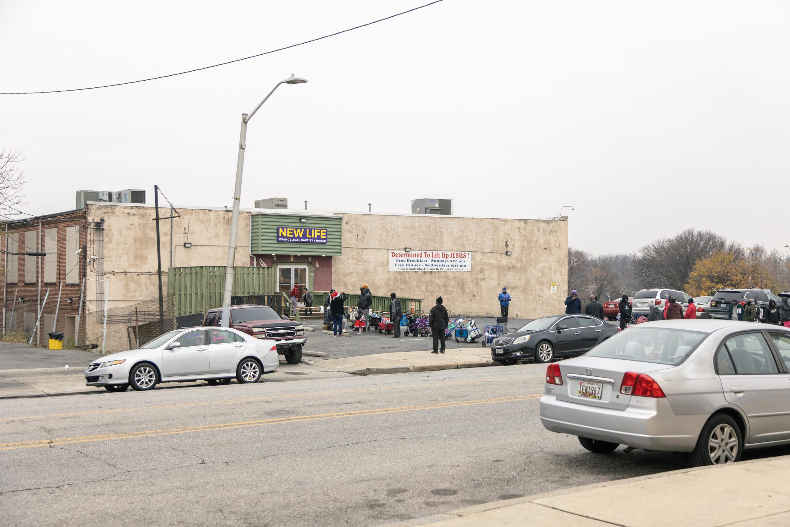 Crowd gathered outside the New Life Evangelical Baptist Church, with individuals waiting in line for free meals and services, set against a cloudy urban backdrop.