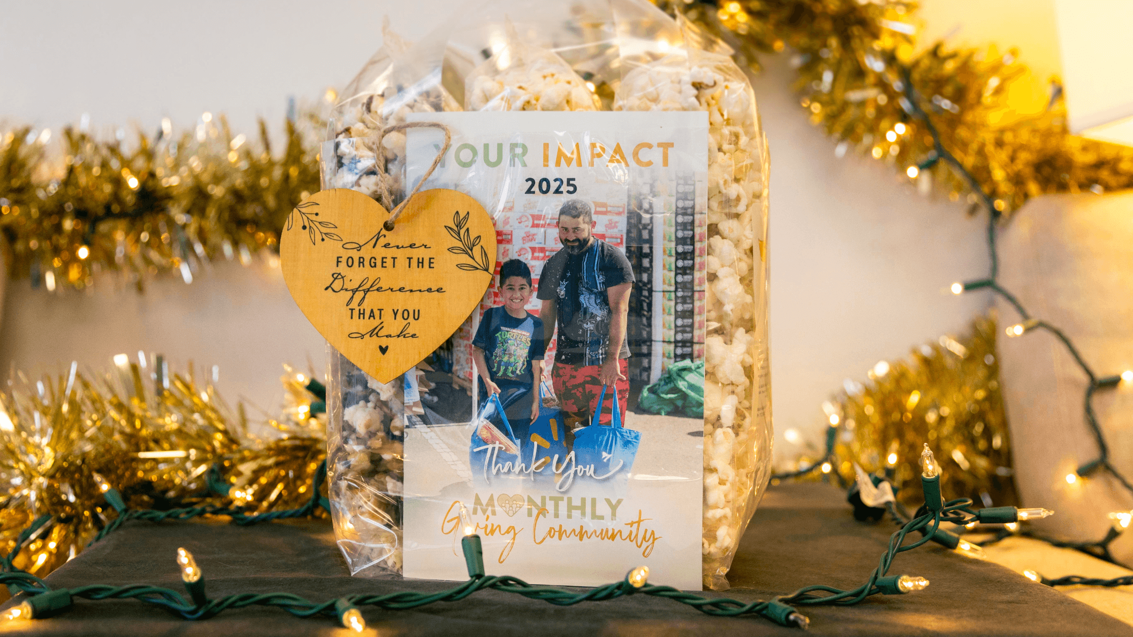 Gift package featuring a thank you card for a monthly giving community, with a photo of a man and a child, and popcorn inside a clear bag, set against a festive background with holiday lights and decorations.