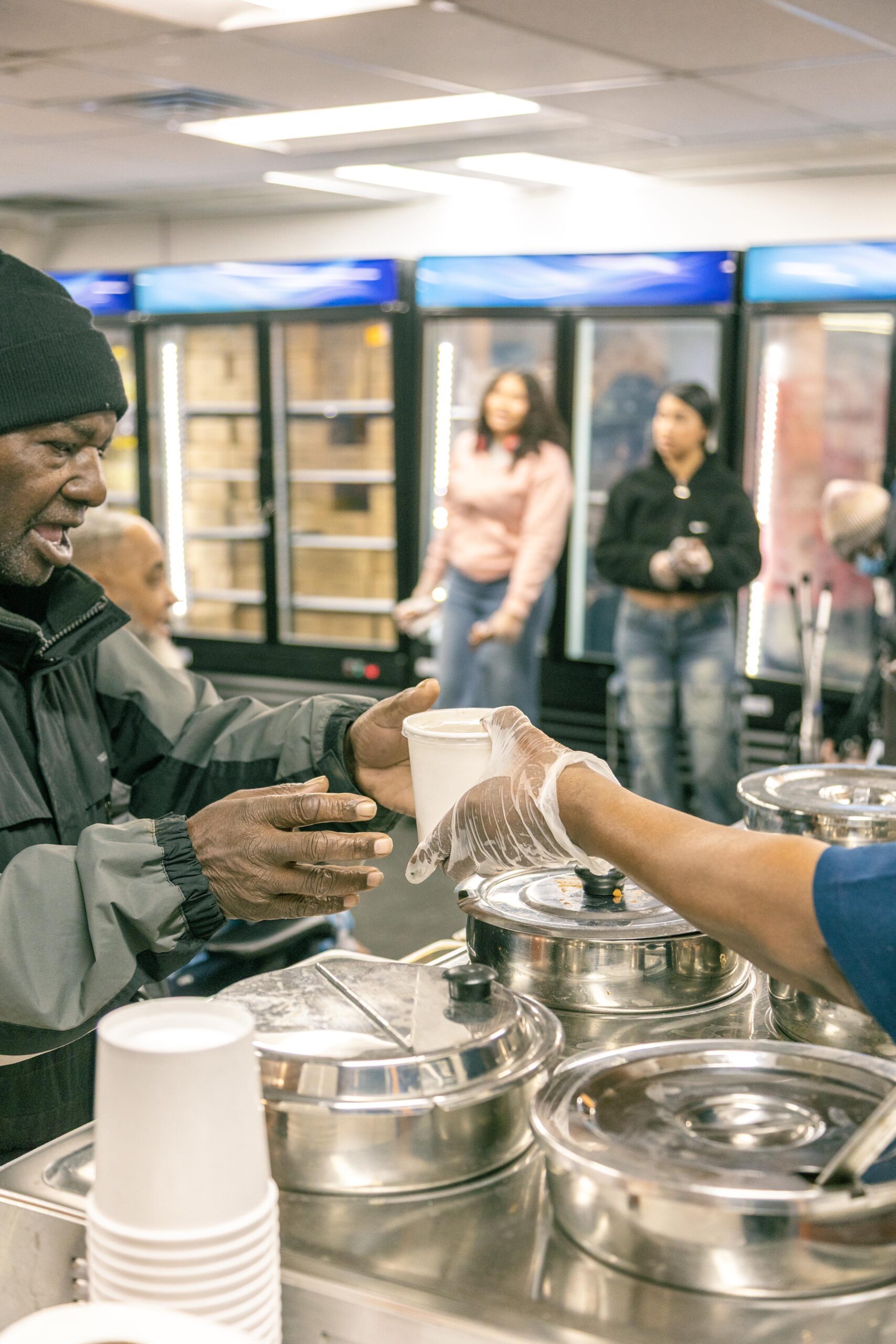 A man in a black beanie receives a food container from a server wearing gloves at a community food distribution event, with people waiting in the background.