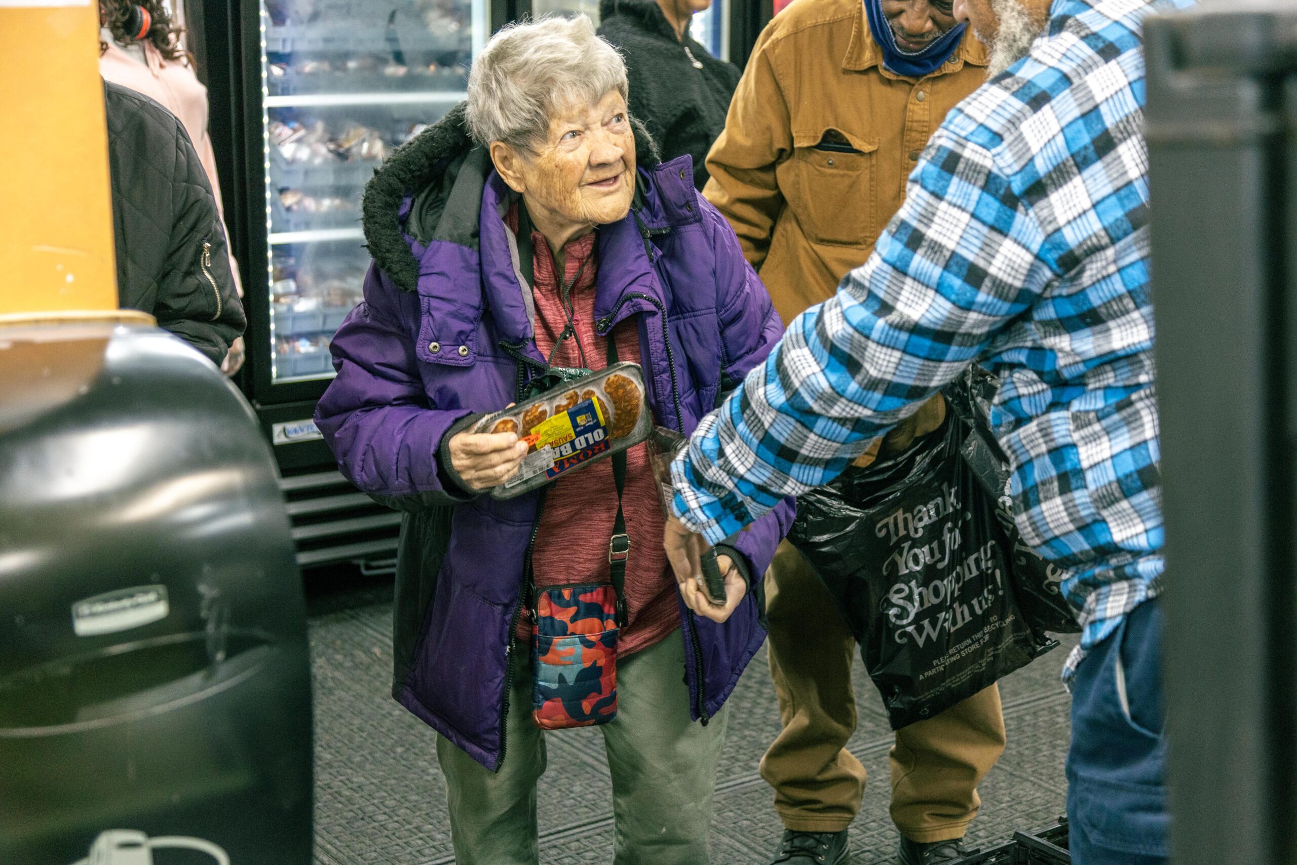 An elderly woman in a purple jacket smiles while holding a food package in a busy grocery store, engaging with a man in a plaid shirt. The background features shelves and other shoppers, creating a lively shopping atmosphere.