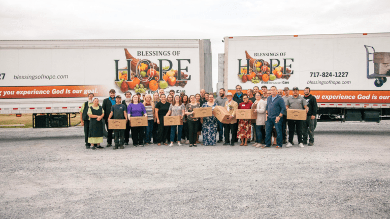 Group photo of volunteers and staff from Blessings of Hope, standing in front of two food distribution trucks, holding cardboard boxes filled with food. The image highlights community support and the mission to provide hope and nourishment.