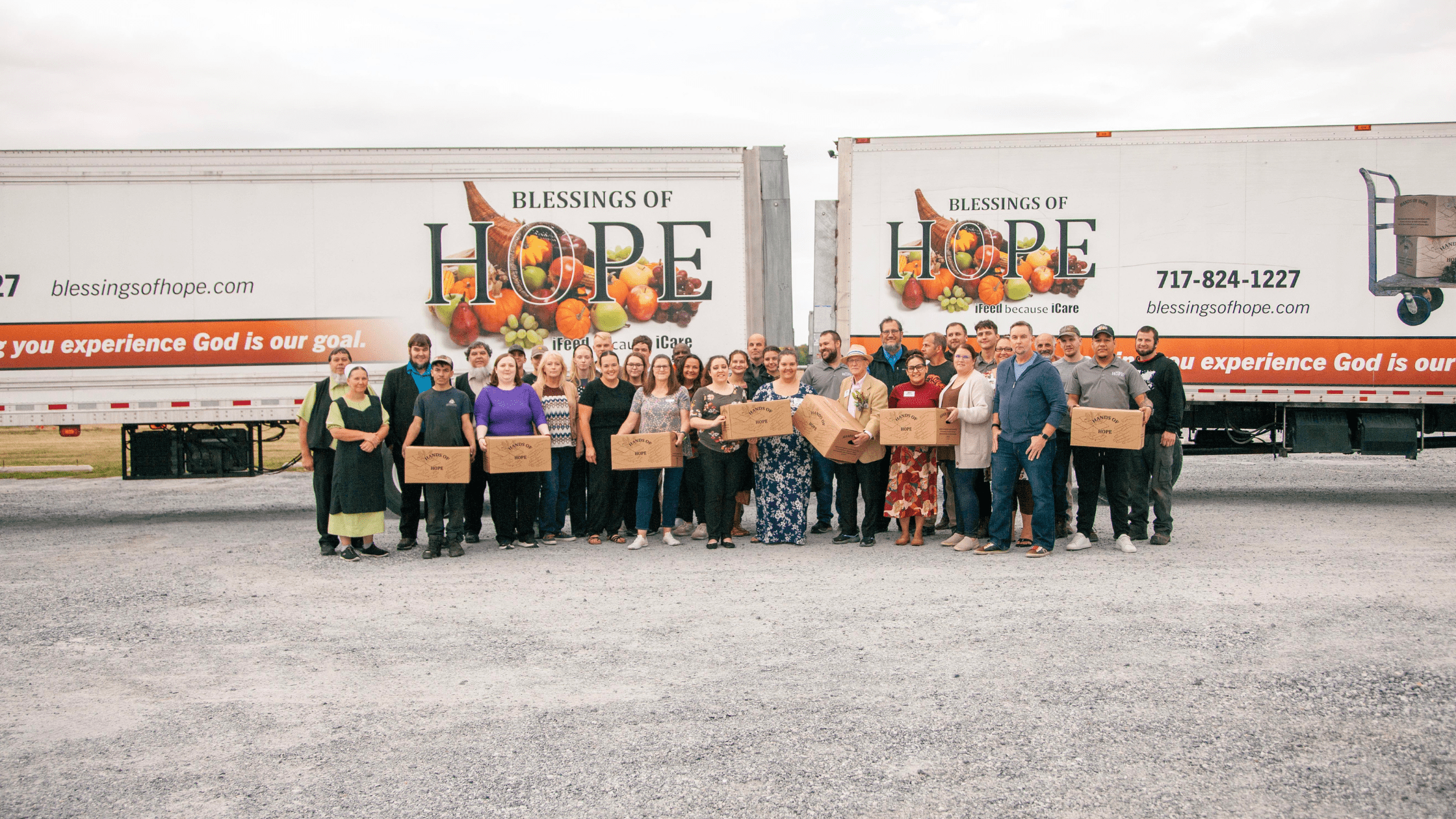 Group photo of volunteers and staff from Blessings of Hope, standing in front of two food distribution trucks, holding cardboard boxes filled with food. The image highlights community support and the mission to provide hope and nourishment.