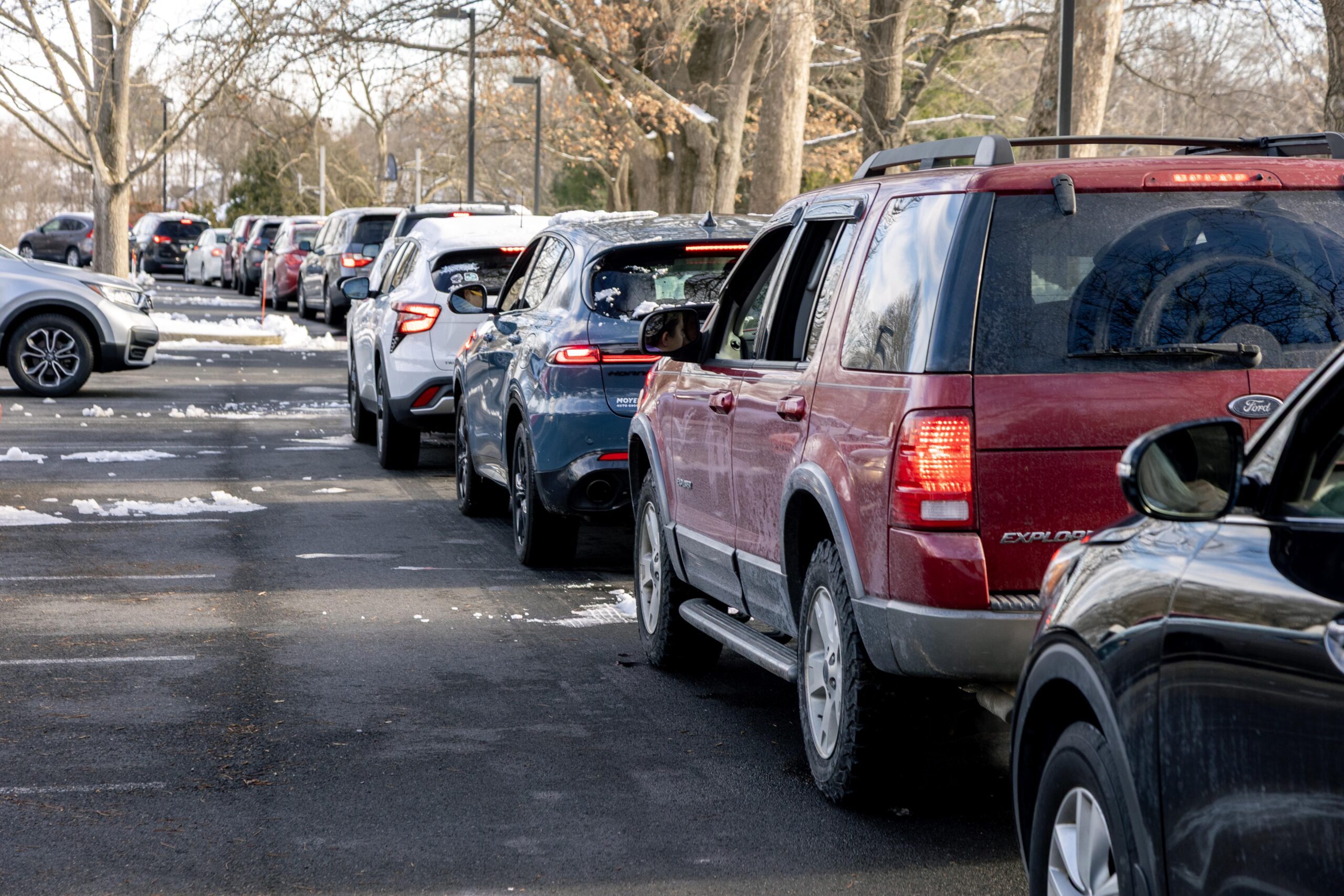 Line of vehicles waiting in a snowy parking lot, featuring a mix of SUVs and cars, with trees in the background.