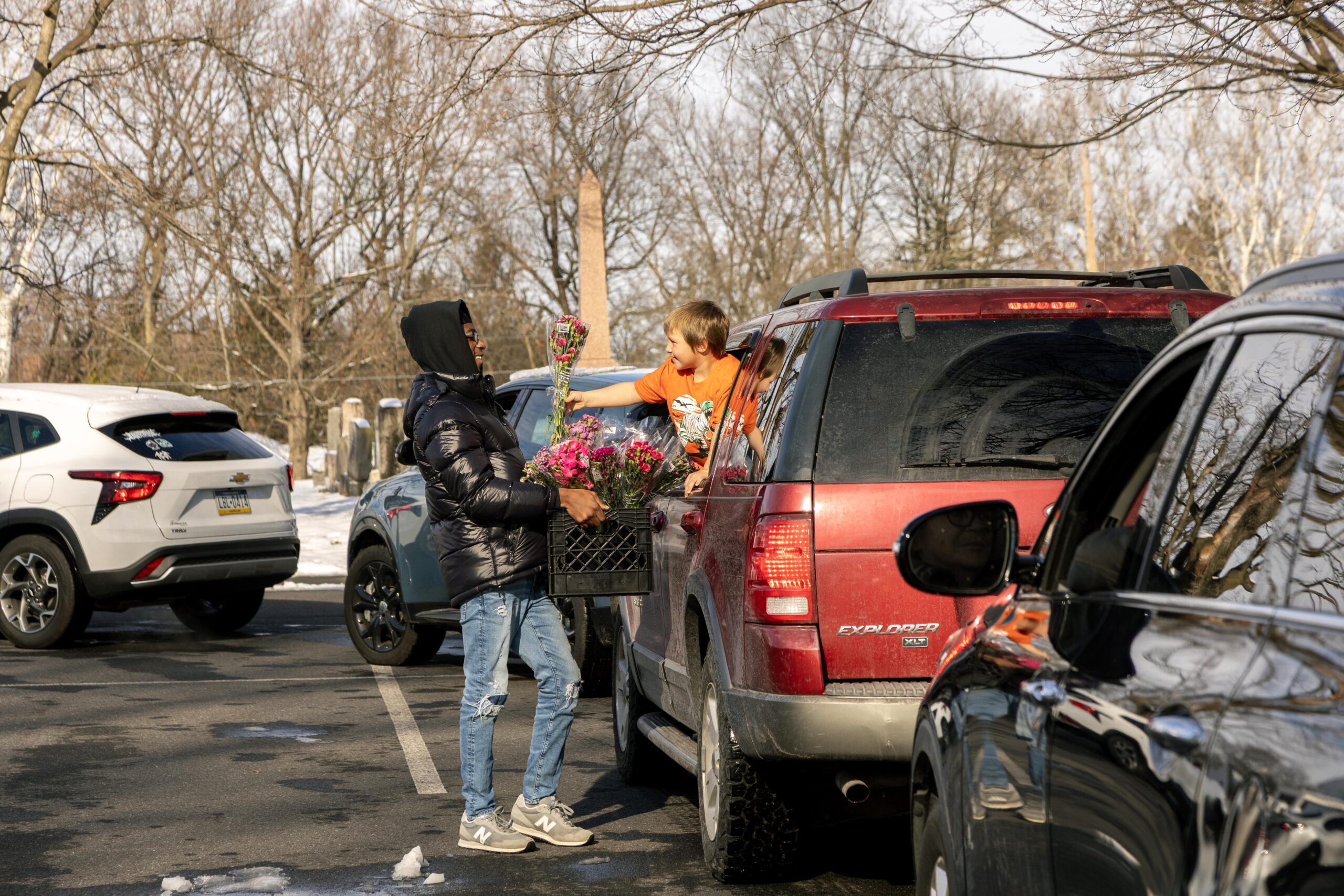 A young boy in an orange shirt hands a bouquet of flowers to a man in a black puffer jacket outside a parked red SUV, surrounded by winter trees and other vehicles in a parking lot.