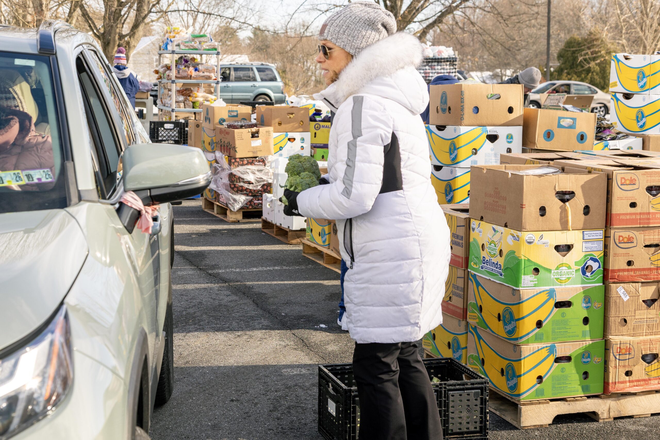 Woman in a white winter coat holding broccoli at a food distribution event, with boxes of fruits and vegetables in the background and a car nearby.