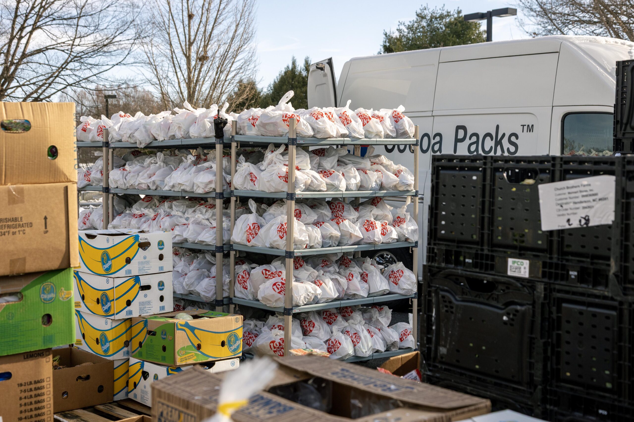 Shelves filled with white bags labeled with red hearts, stacked outside near a delivery van. Boxes of bananas and other produce are scattered around, indicating a food distribution or donation effort.