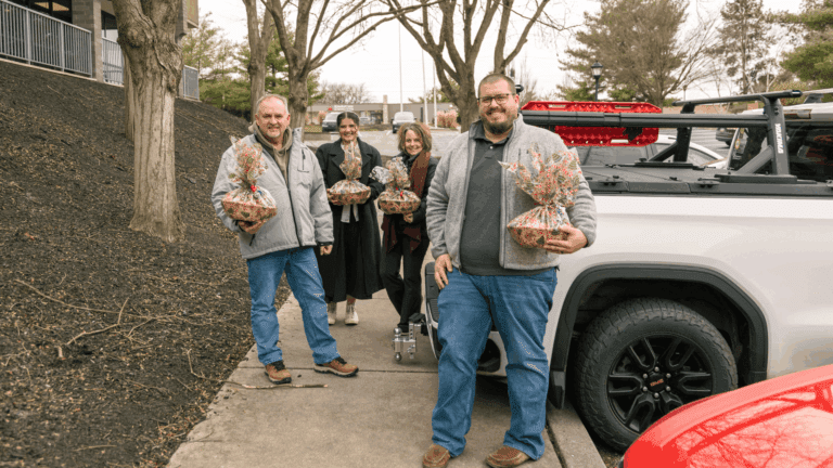 Group of four people standing outside a building, each holding a decorative gift basket. They are smiling and dressed in casual winter attire, with a pickup truck parked nearby. Trees line the walkway, creating a pleasant outdoor setting.