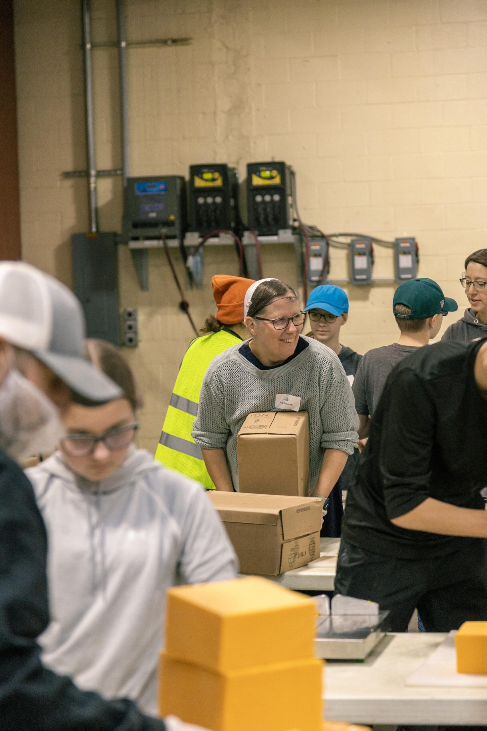 Volunteers packing food boxes in a warehouse, with a focus on a smiling woman carrying a box. The scene highlights teamwork and community involvement in food distribution efforts.