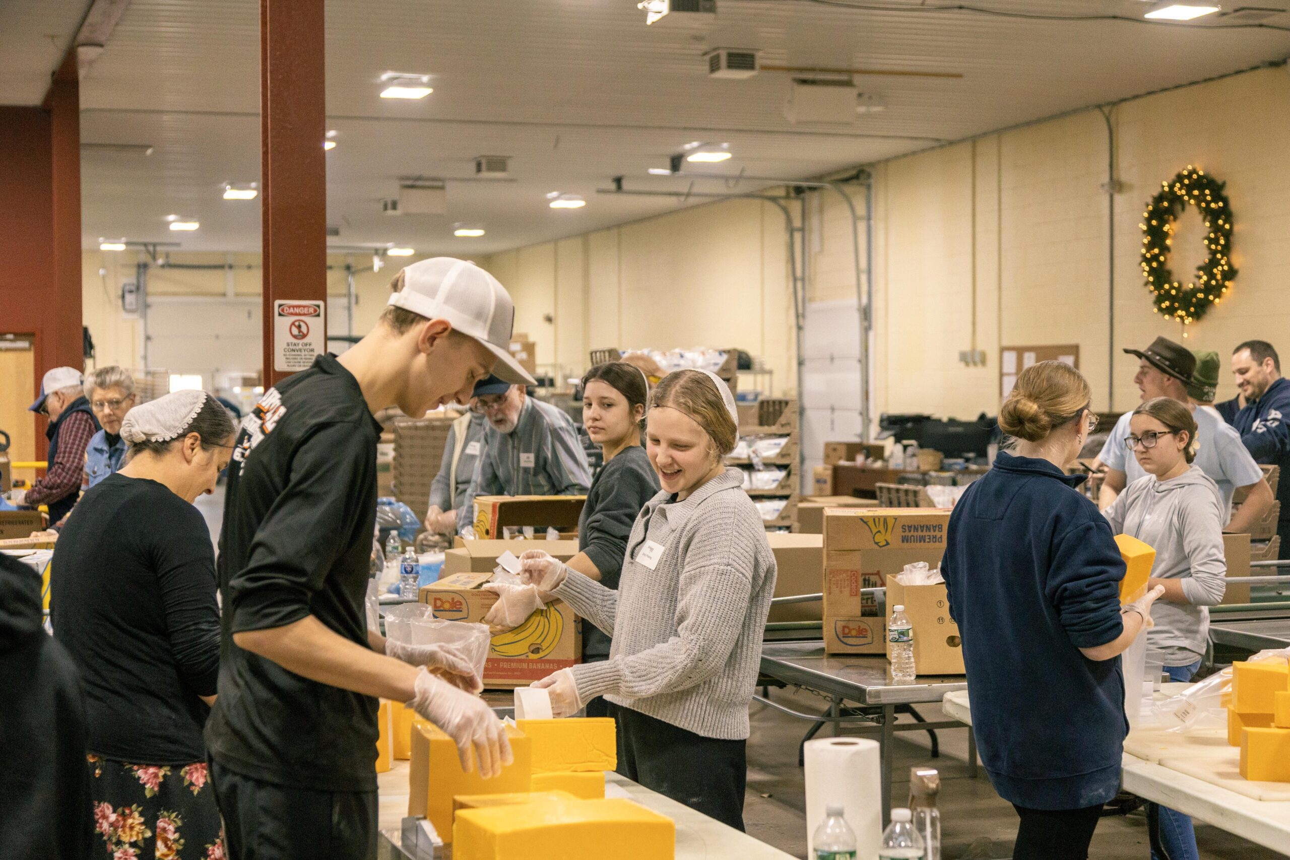 Volunteers engaged in food packaging at a community event, working together in a warehouse setting with yellow boxes and various supplies visible on tables.