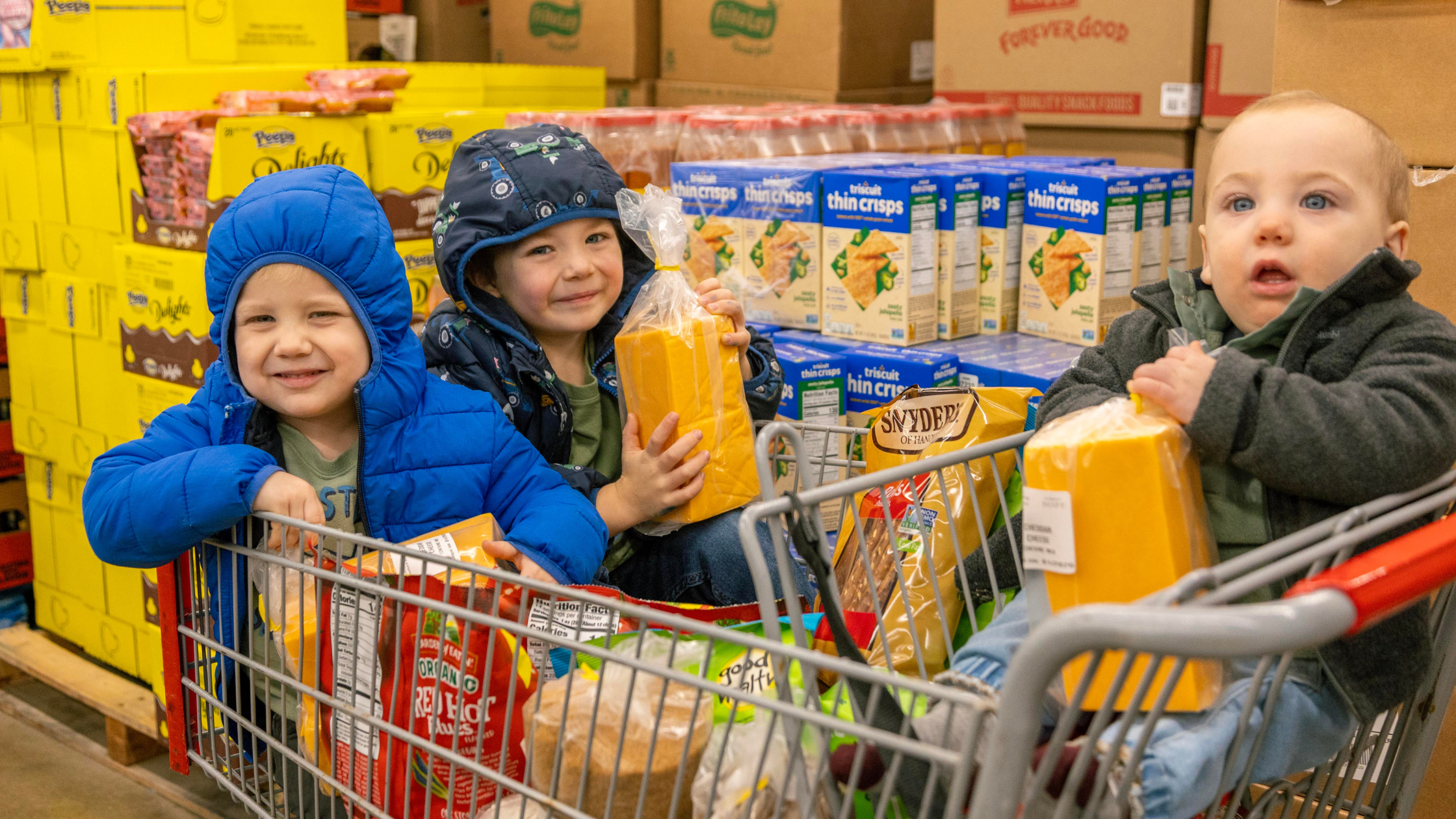 Three children smiling in a shopping cart filled with snacks and groceries, surrounded by colorful packaged goods in a store aisle. The boys are wearing winter jackets and holding blocks of cheese, showcasing a fun family shopping experience.