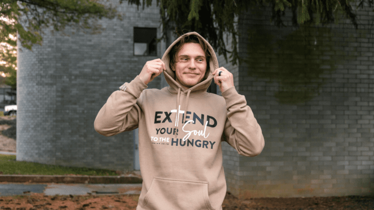 Young man wearing a beige hoodie with the message "Extend Your Soul to the Hungry" stands outdoors in front of a gray brick building, smiling and adjusting his hood. The image conveys a sense of warmth and community engagement.