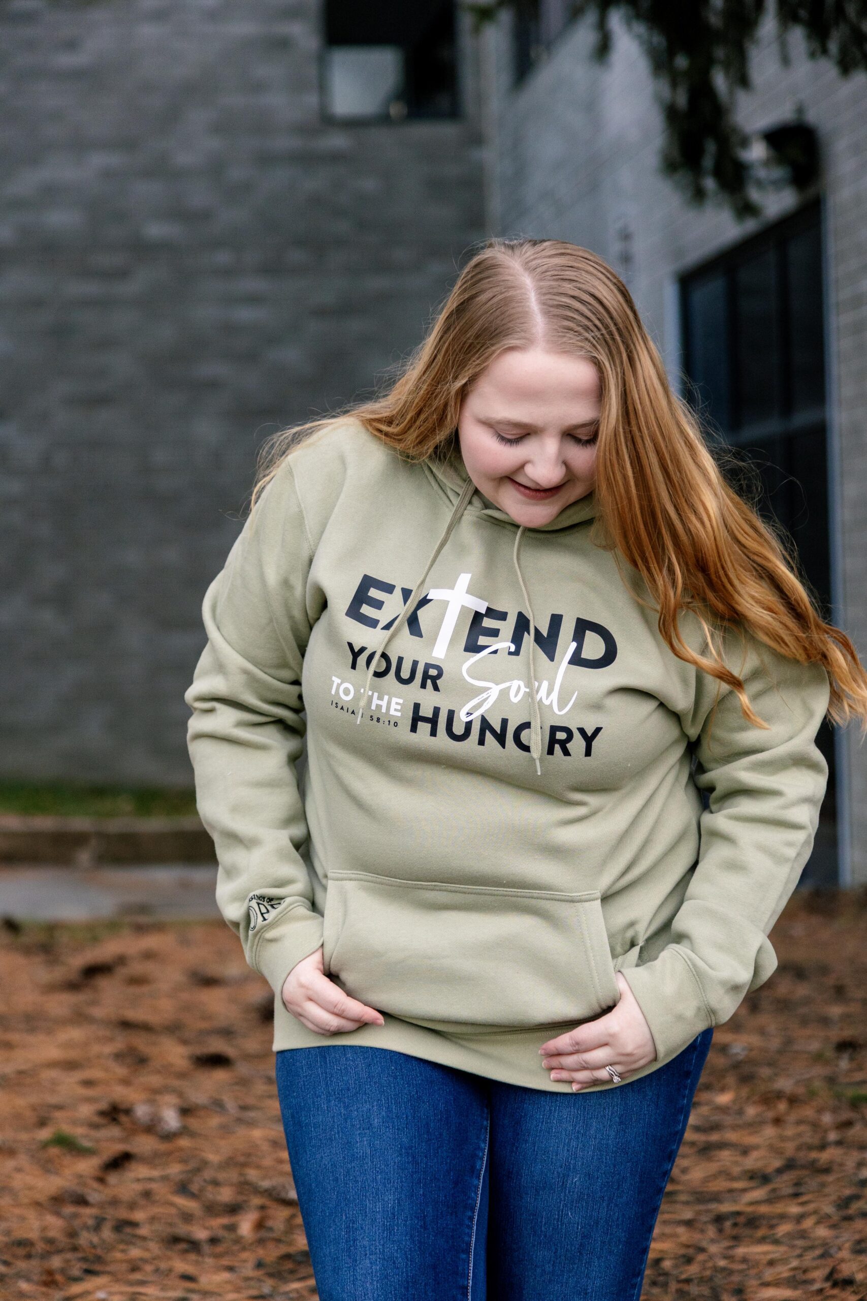 Young woman wearing a green hoodie with the text "Extend Your Soul to the Hungry" in a casual outdoor setting, smiling and looking down. The background features a modern building and natural elements.