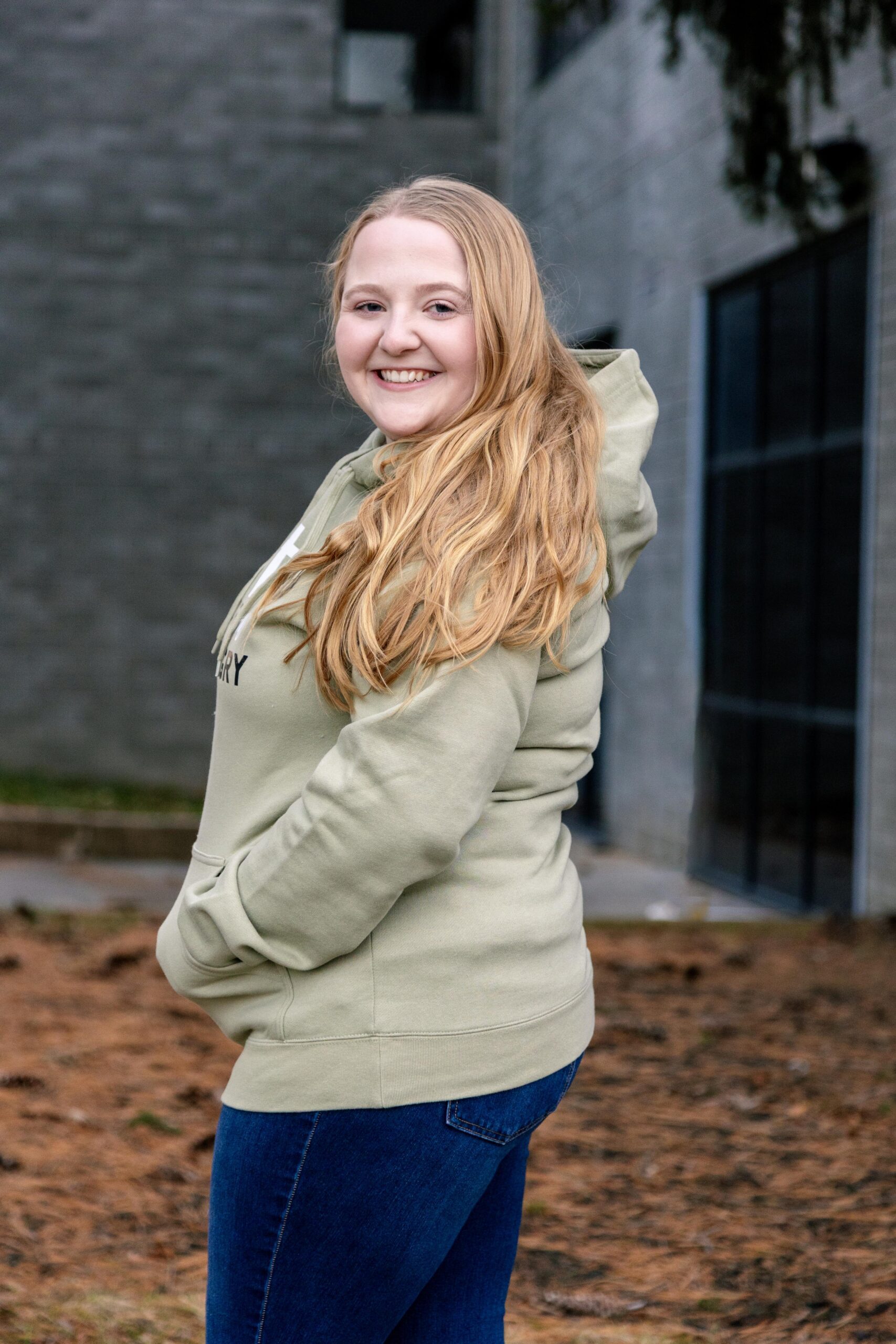 Young woman smiling while wearing a light green hoodie and blue jeans, standing outdoors in front of a modern building. The background features a natural setting with pine needles on the ground.