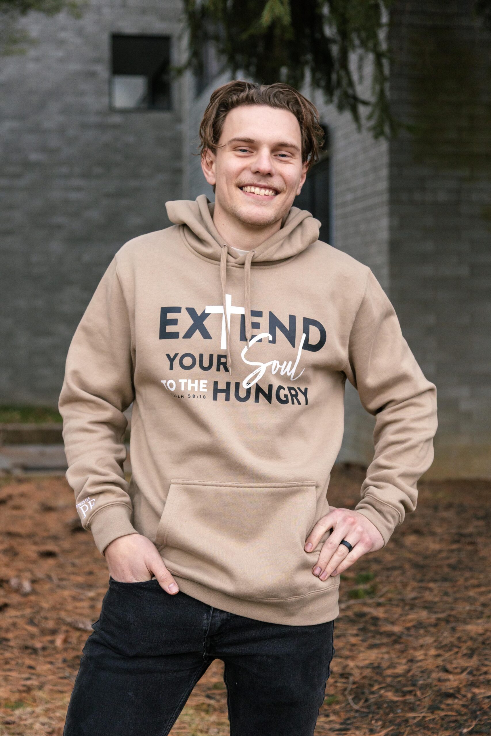 Young man smiling while wearing a beige hoodie with the text "Extend Your Soul to the Hungry" prominently displayed. The background features a natural setting with pine needles on the ground and a blurred building in the distance, conveying a casual and inviting atmosphere.