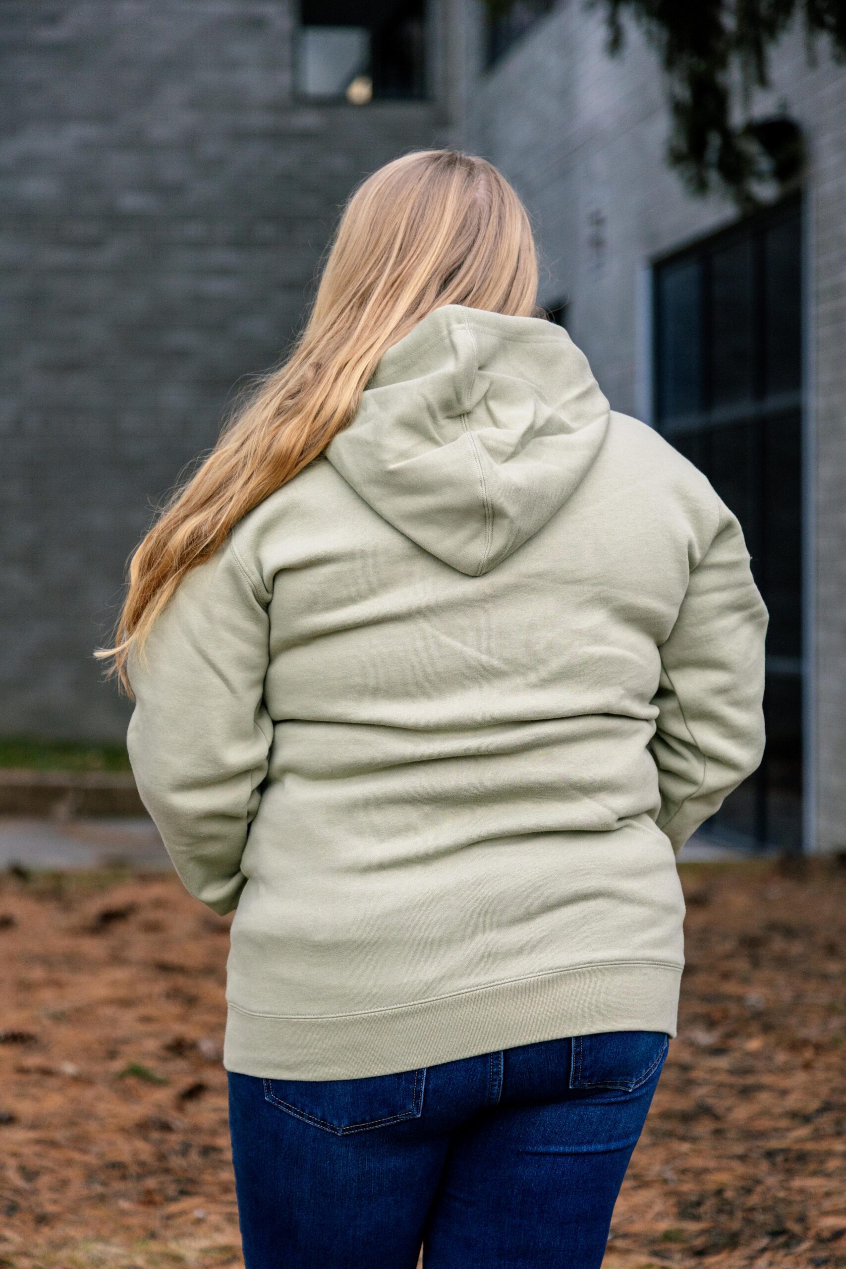 Woman wearing a light green hoodie and blue jeans, standing outdoors with her back to the camera, against a backdrop of a modern building and natural landscape.