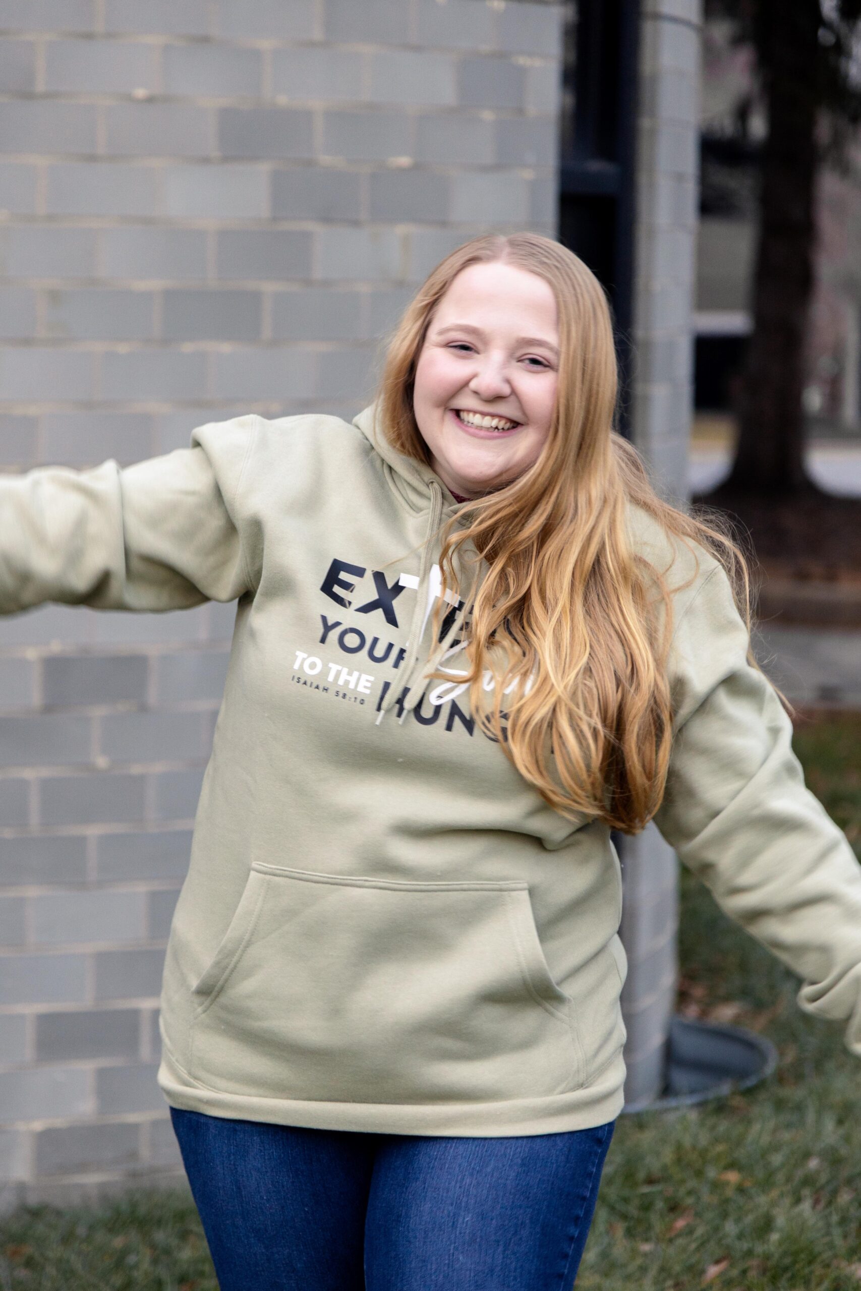 Smiling young woman with long blonde hair wearing a light green hoodie and blue jeans, joyfully posing outdoors in front of a gray brick wall.