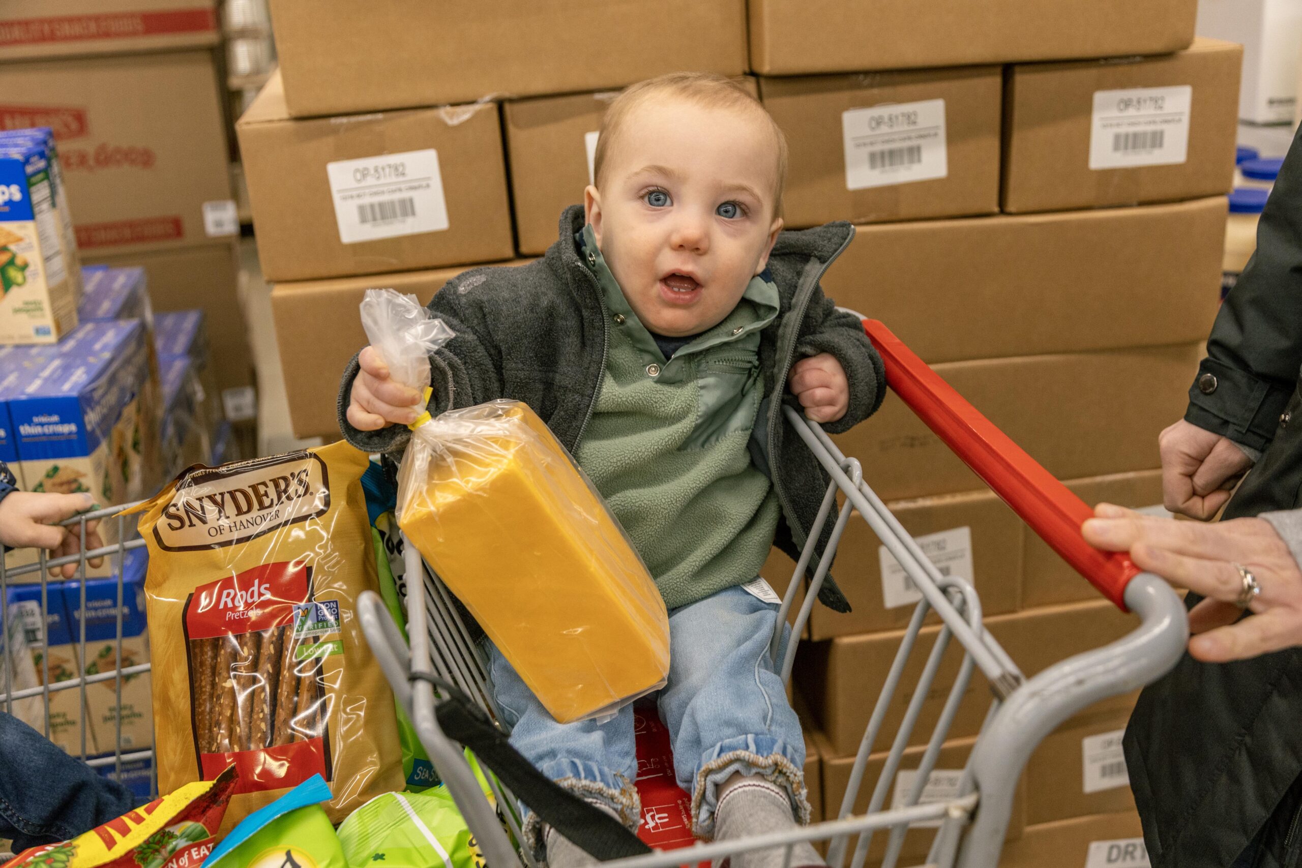 Toddler sitting in a shopping cart holding a block of cheese and surrounded by snacks, with boxes stacked in the background at a grocery store.
