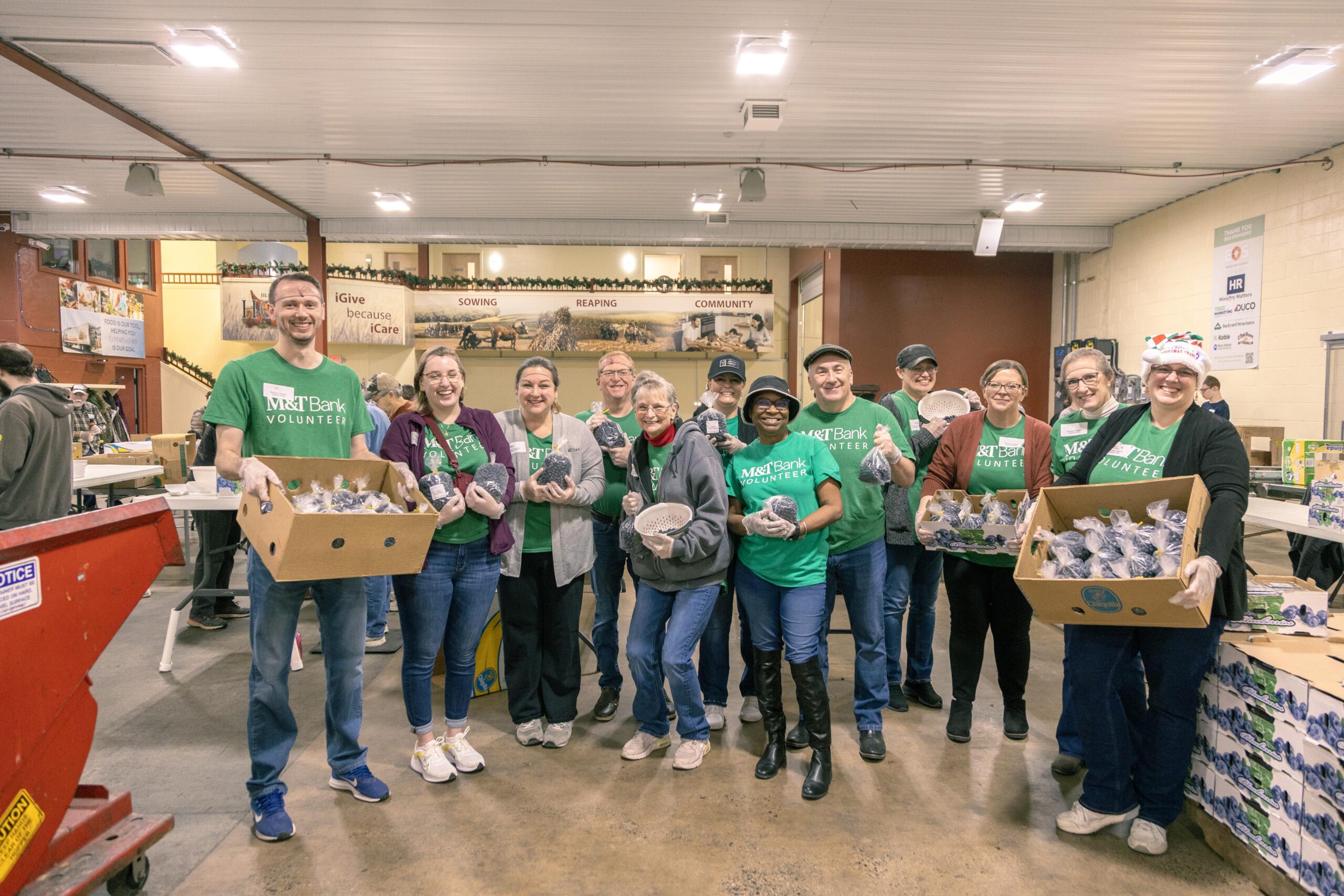 Group of M&T Bank volunteers smiling and holding boxes of food items in a community service event, inside a spacious facility decorated for the holiday season.