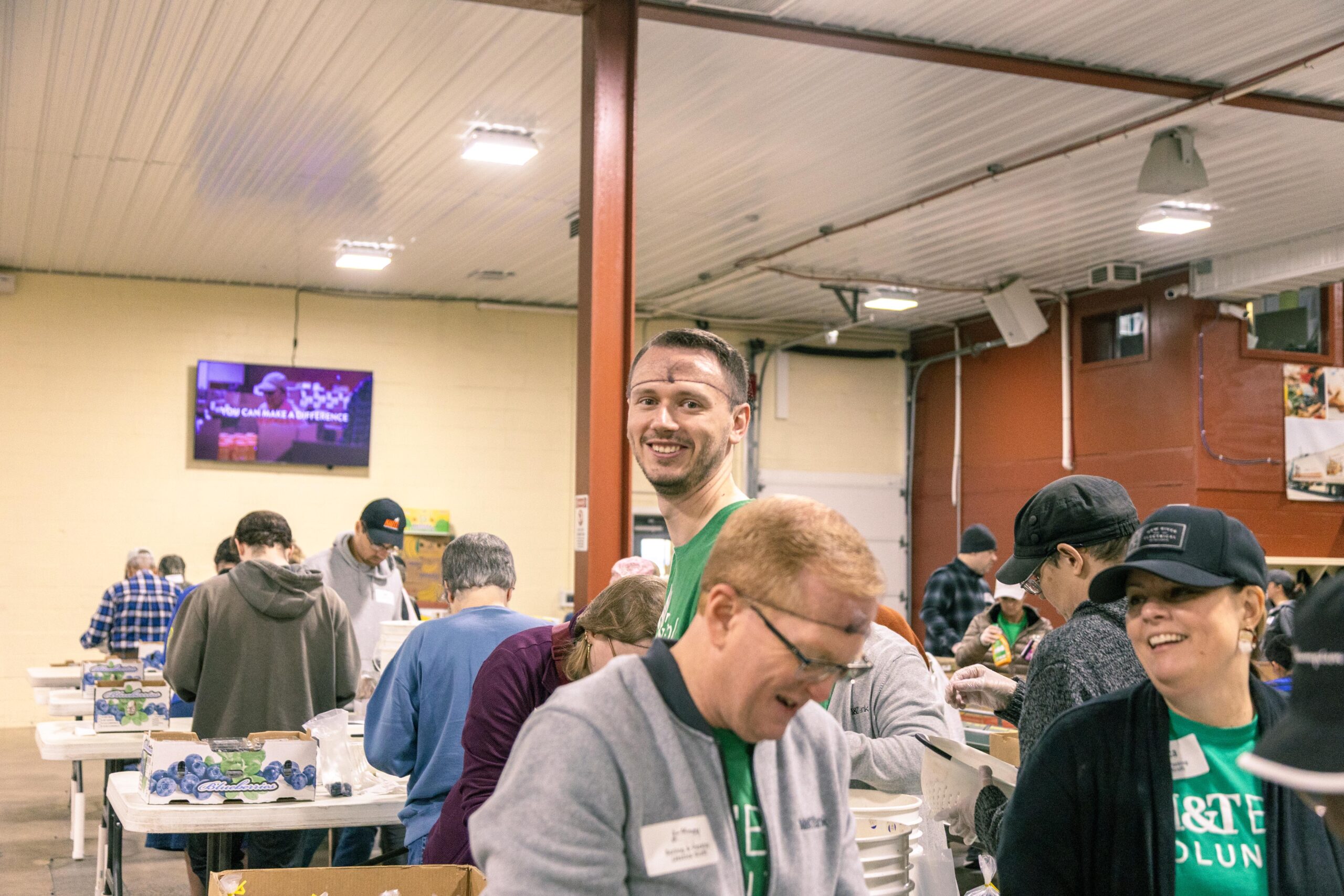 Volunteers sorting food in a community center, smiling and engaging in teamwork, with a television screen displaying a motivational message in the background.