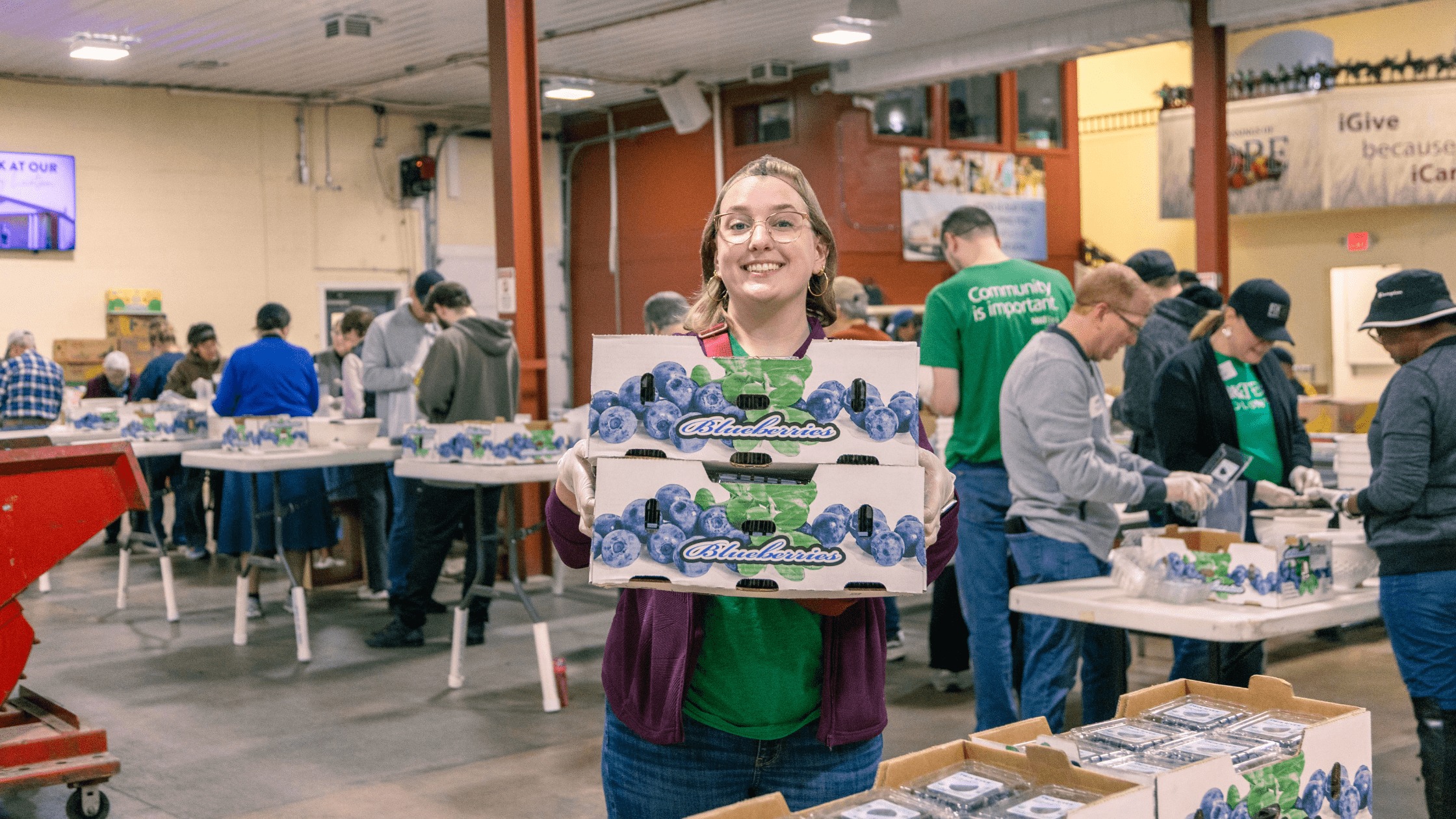 Volunteer smiling while holding boxes of blueberries at a community food distribution event, with other volunteers sorting food in the background.