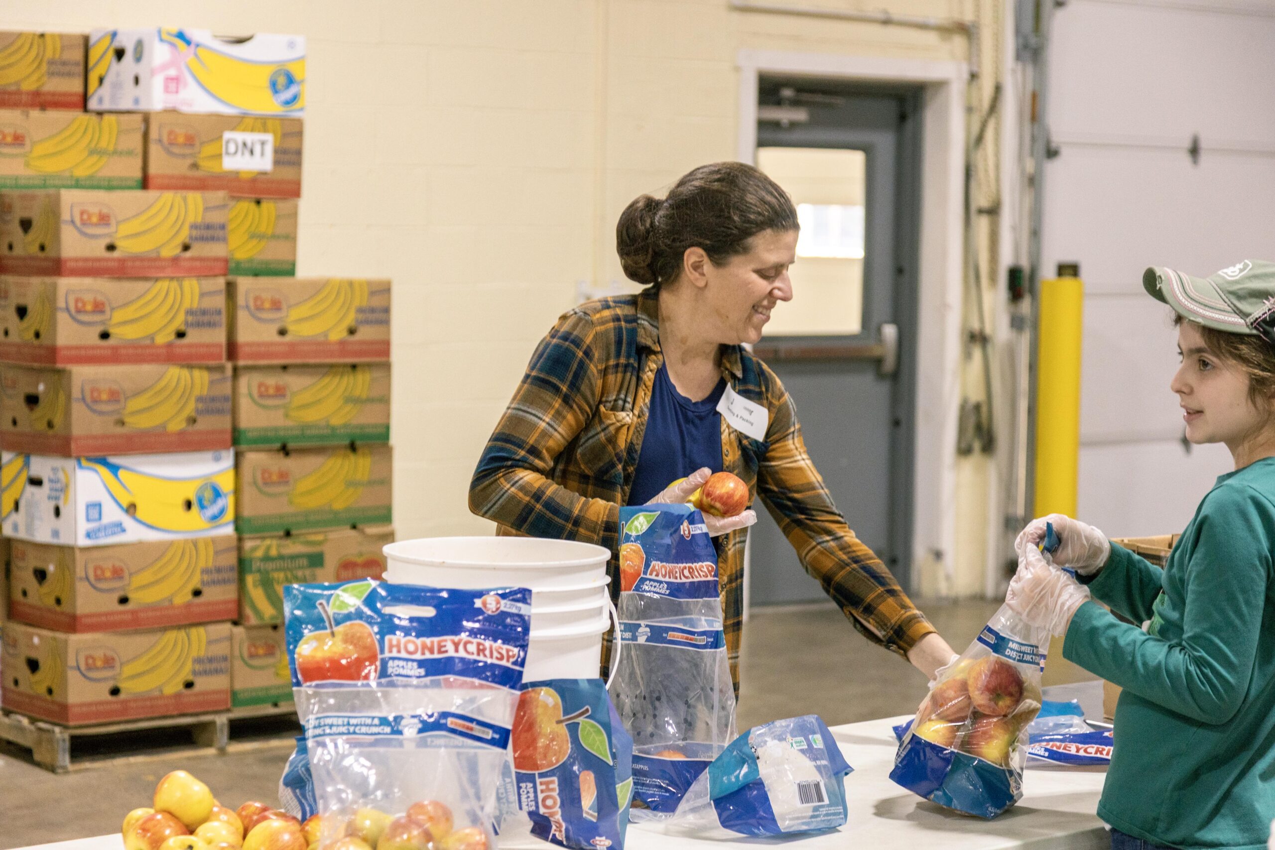 Volunteers packaging Honeycrisp apples at a food distribution center, with boxes of bananas stacked in the background.