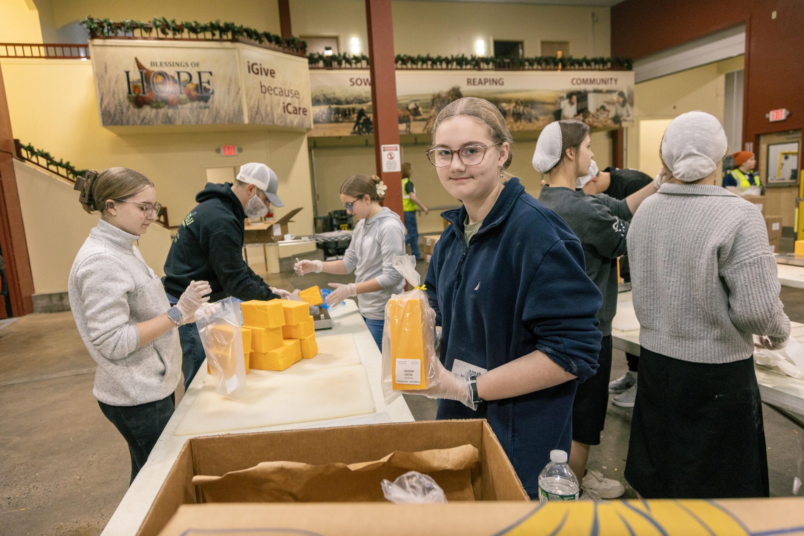Volunteers packaging food items at a community service event, with a focus on a young woman holding a bag of cheese. The setting includes a large banner displaying "Blessings of Hope" and other volunteers working in the background.