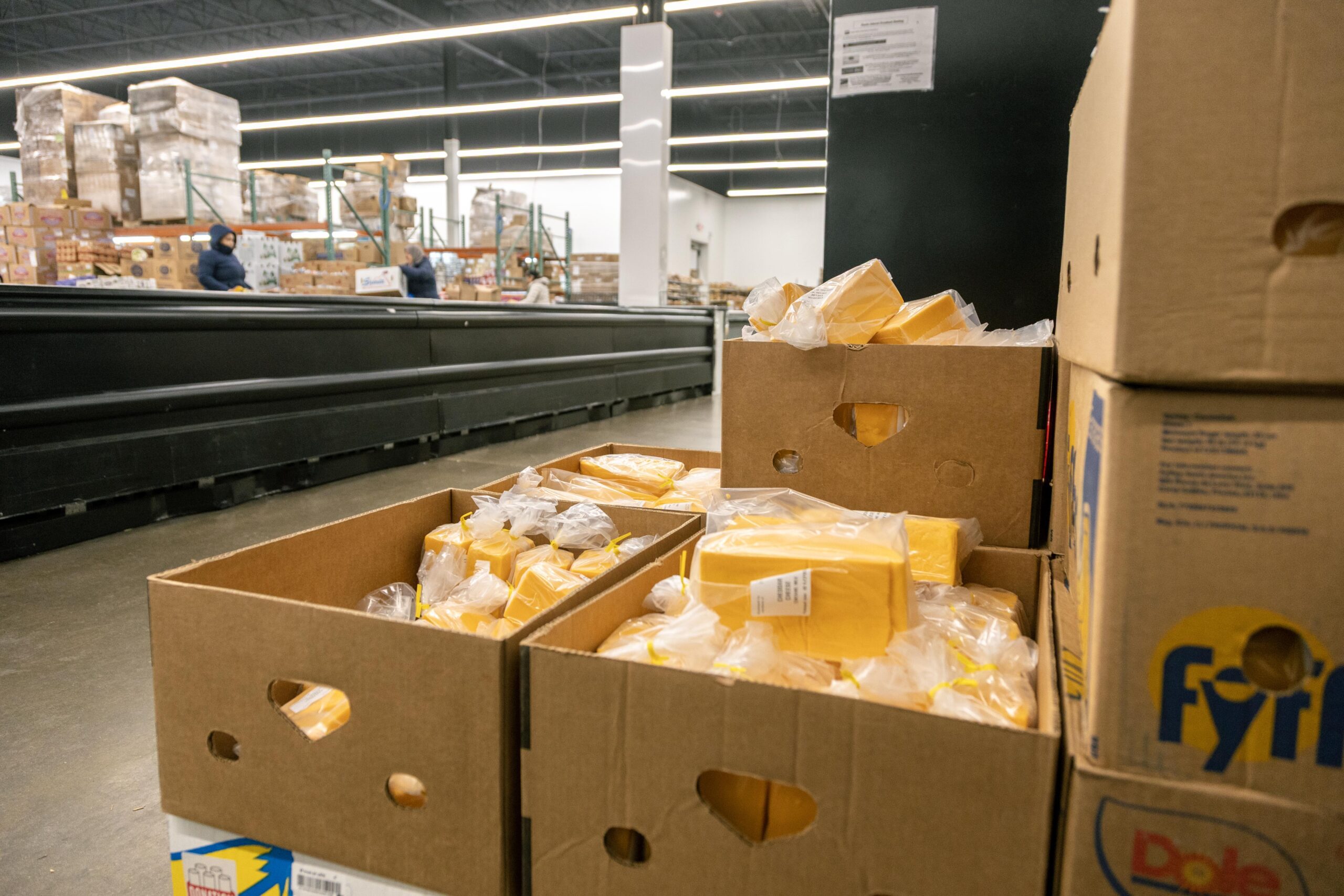 Boxes filled with individually packaged yellow cheese blocks stacked in a warehouse, with shelves of grocery items in the background. Ideal for food distribution and inventory management.