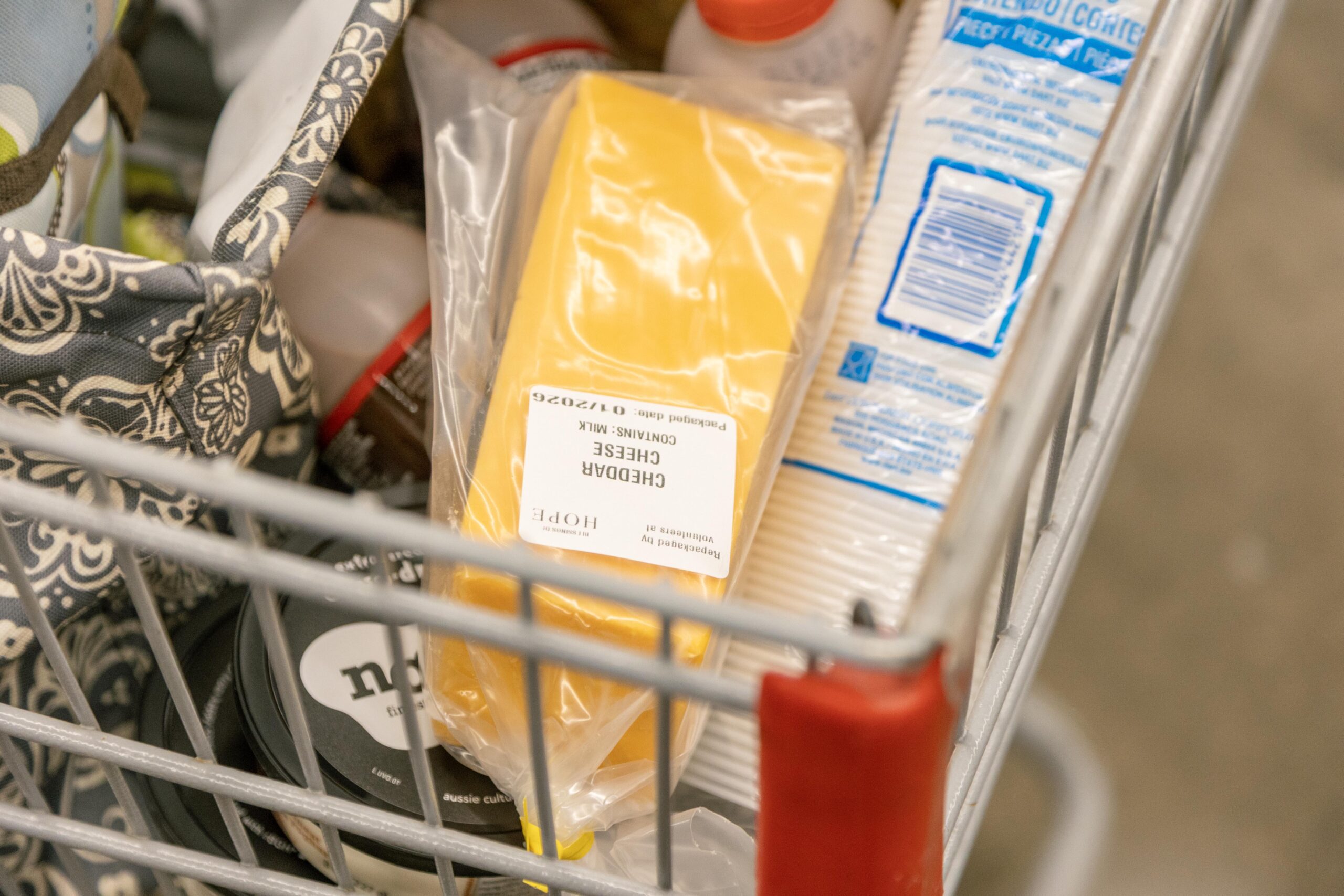 Grocery cart filled with various items, including a package of cheddar cheese, milk, and other food products, showcasing a shopping experience at a supermarket.