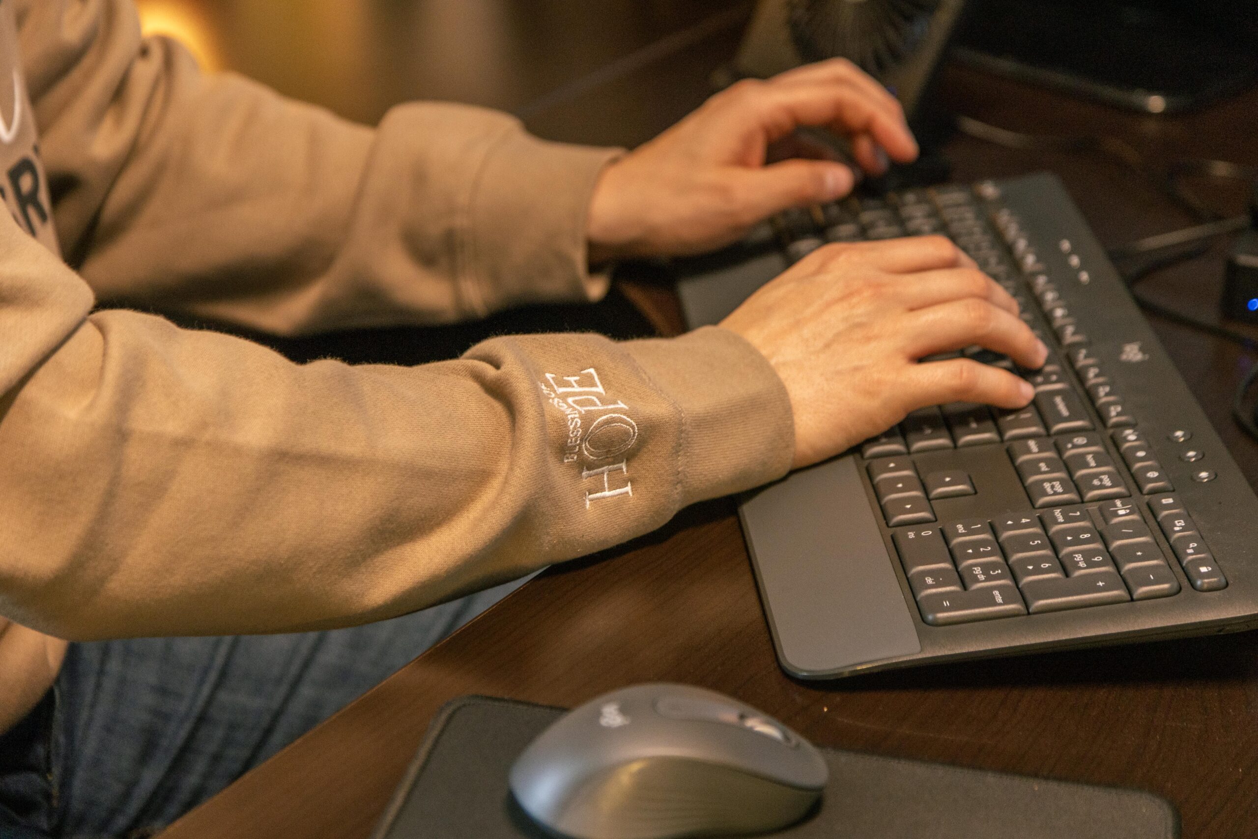 Person typing on a keyboard while wearing a brown sweatshirt, with a computer mouse and a fan visible on the desk.