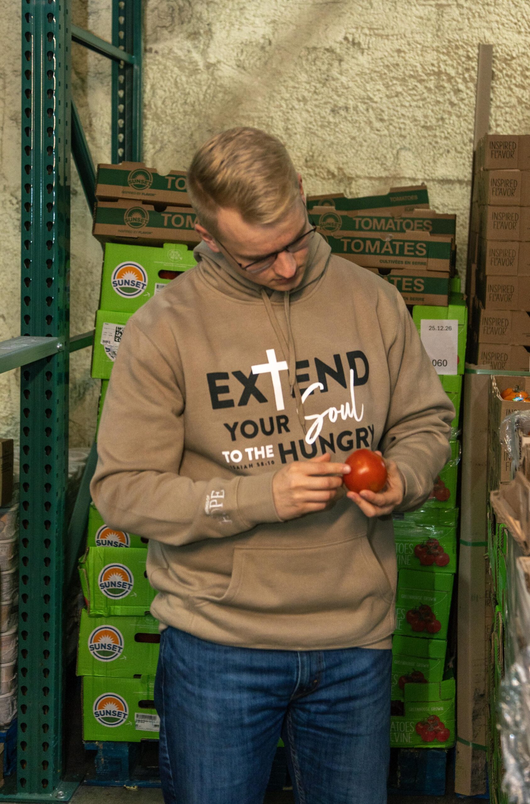 Man in a beige hoodie inspecting a tomato in a food pantry, surrounded by boxes of tomatoes and canned goods, emphasizing community support and food distribution efforts.