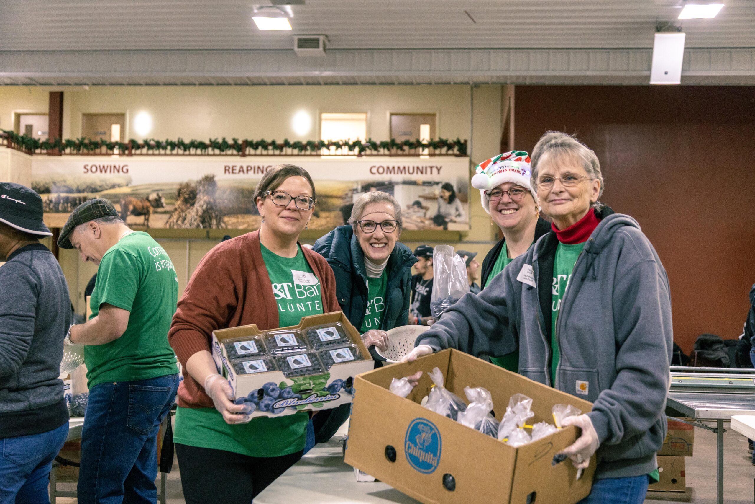 Volunteers smiling and working together at a community food distribution event, holding boxes of fresh produce and packaged goods in a festive environment.