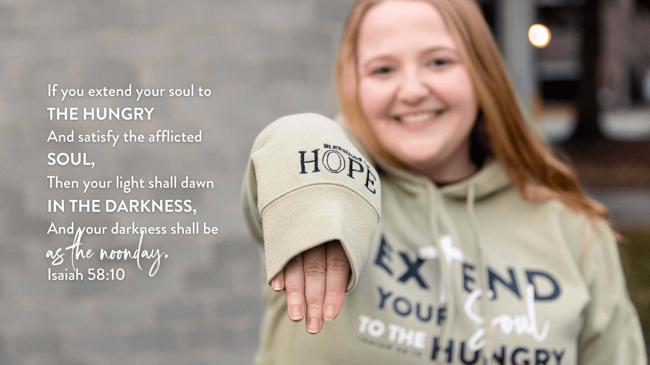 Smiling young woman wearing a hoodie with inspirational text about hope and helping the hungry, with a blurred background. The image emphasizes themes of compassion and community support, featuring a close-up of her hand extending from the oversized sleeve.