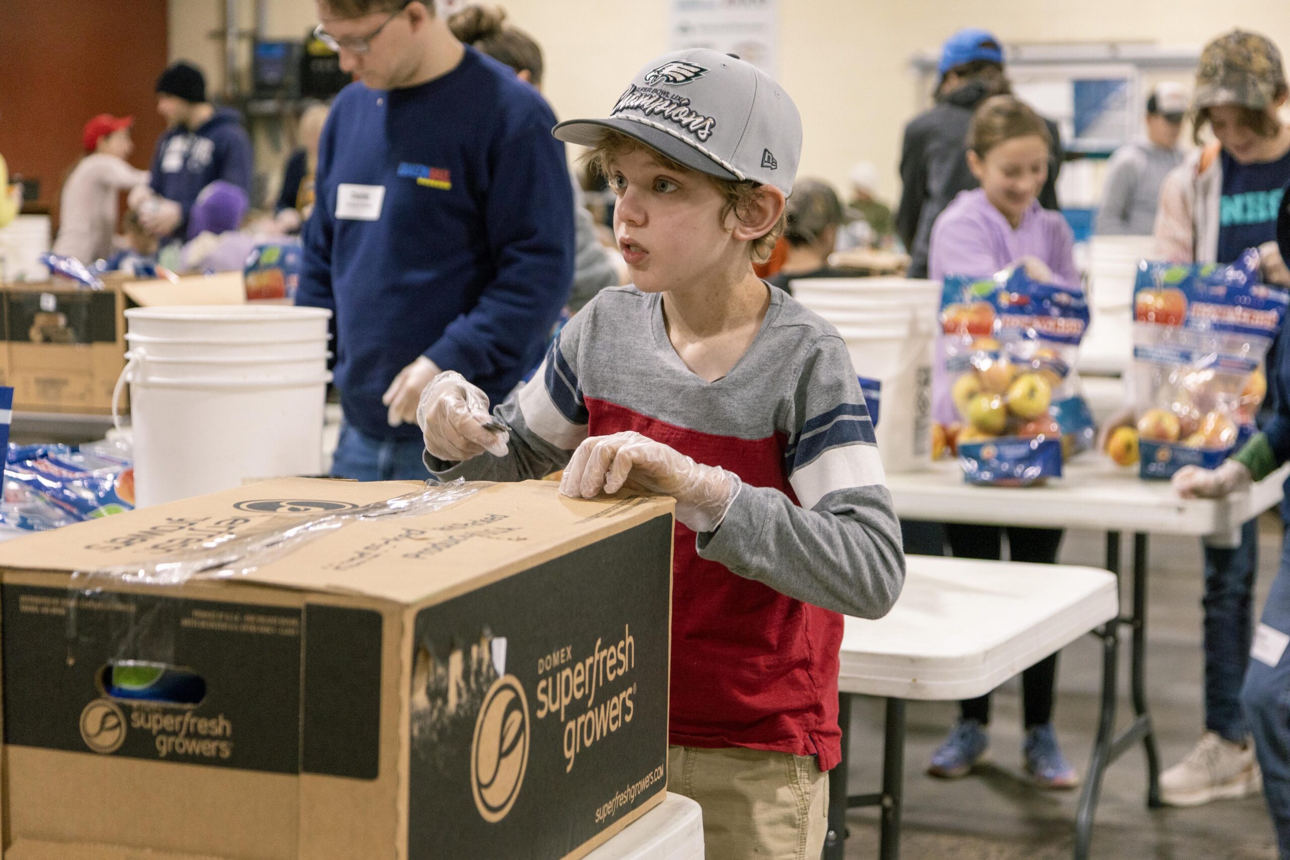 Child volunteering at a food distribution event, carefully sealing a box labeled "superfresh growers" while wearing gloves. Other volunteers are seen in the background, packing bags of apples and organizing supplies on tables.