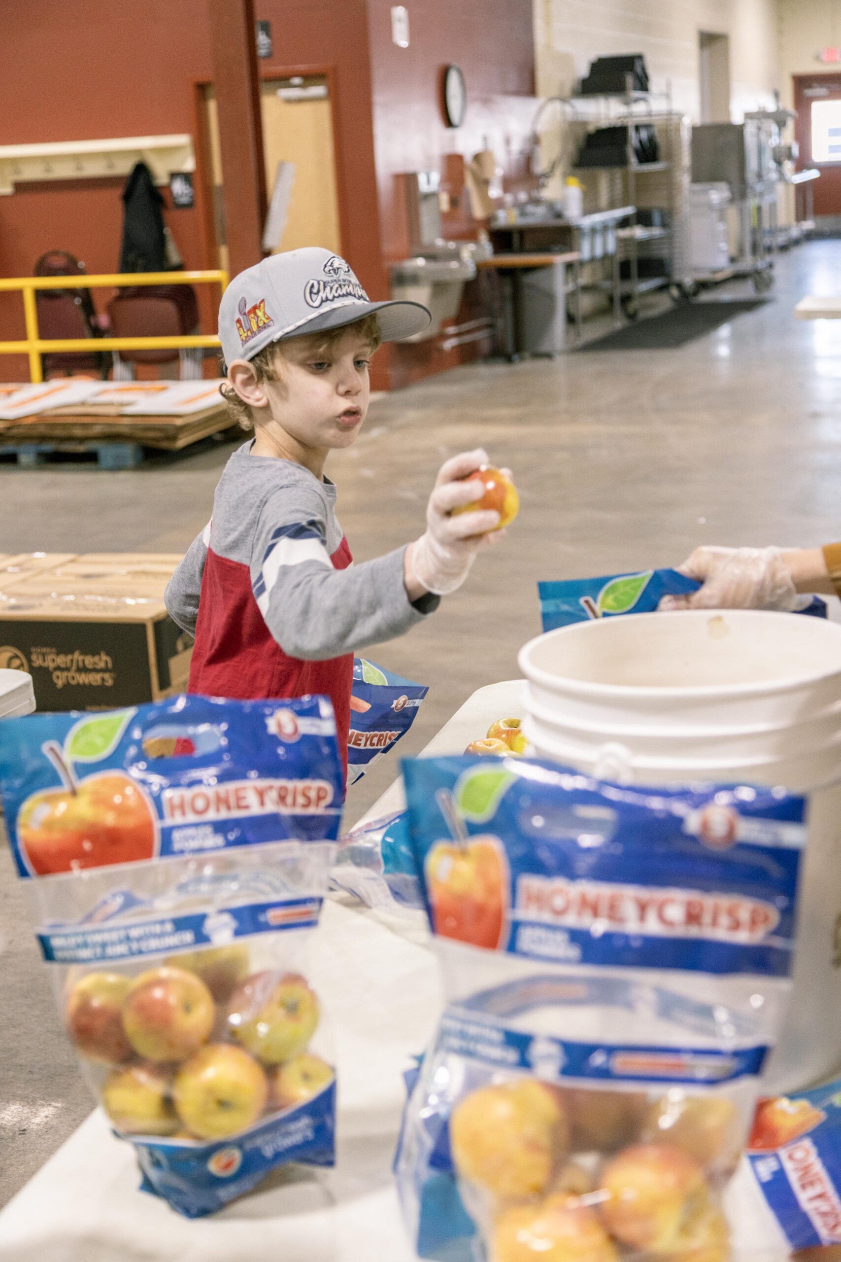 A young boy wearing a baseball cap inspects a Honeycrisp apple while volunteering in a food distribution center, surrounded by bags of apples on a table.
