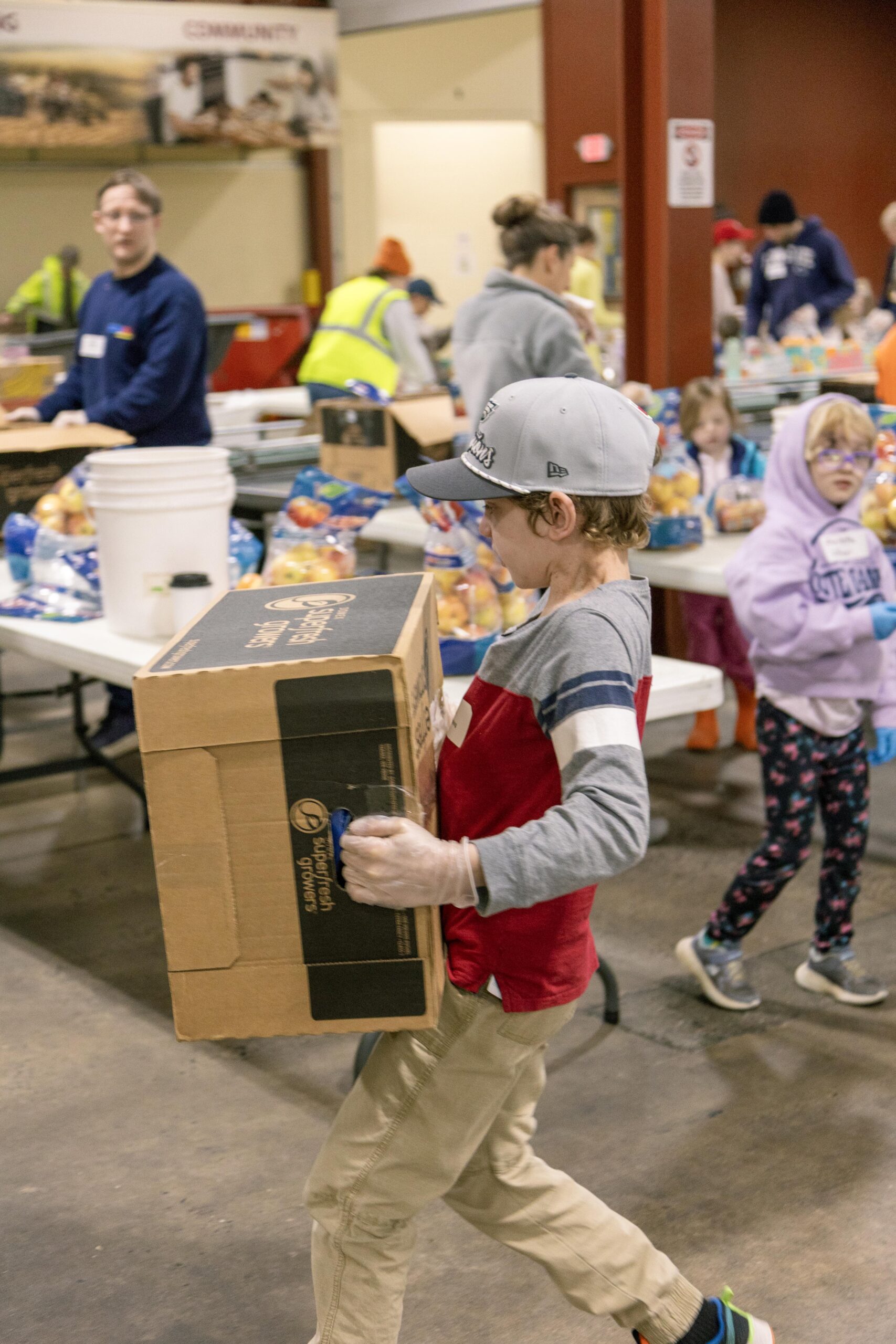 Child carrying a box while volunteering at a food distribution event, surrounded by other volunteers and tables filled with food items.