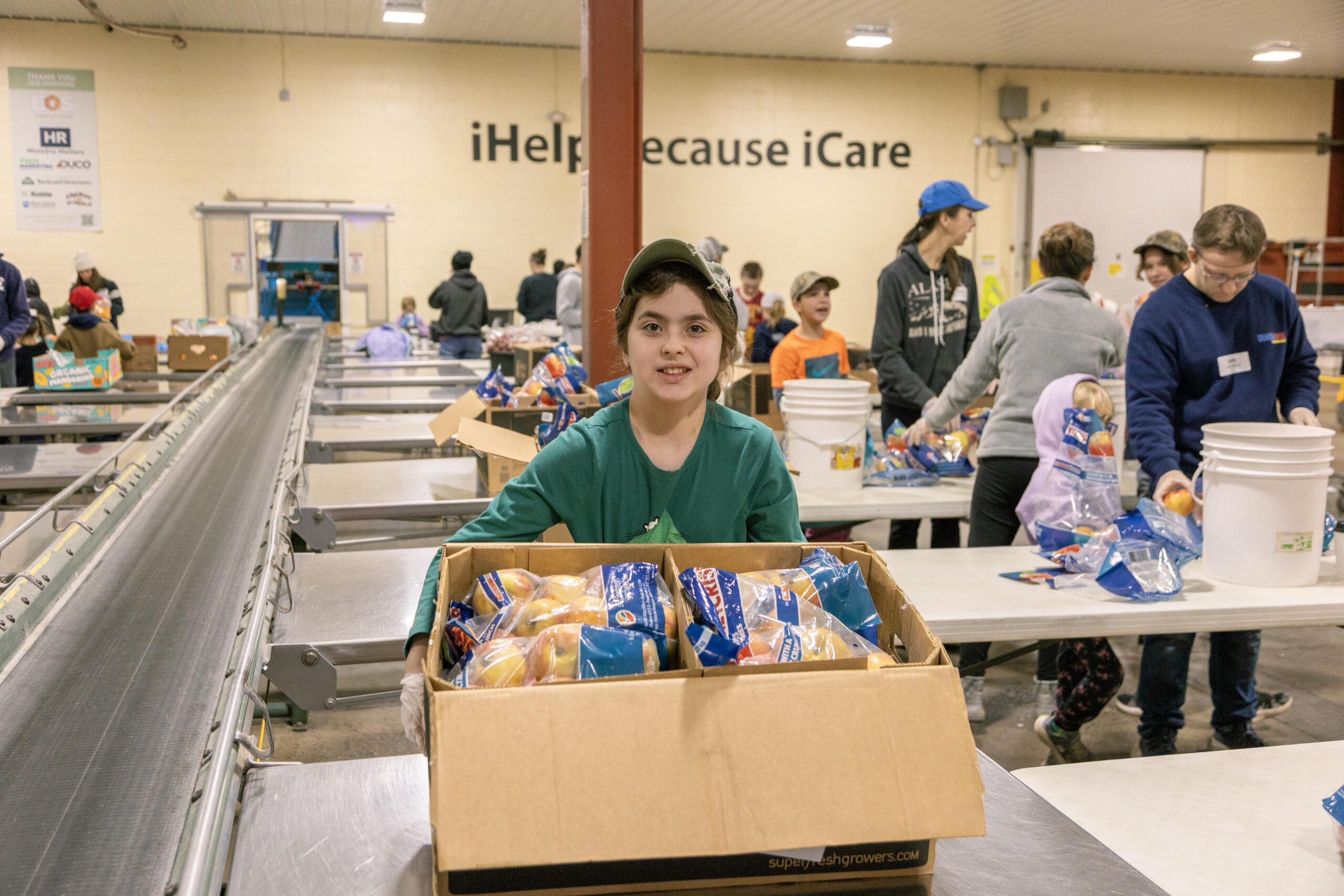 Young volunteer holding a box of packaged apples at a food distribution event, with other volunteers sorting food in the background. The setting features tables and a conveyor belt, highlighting community involvement in food assistance.