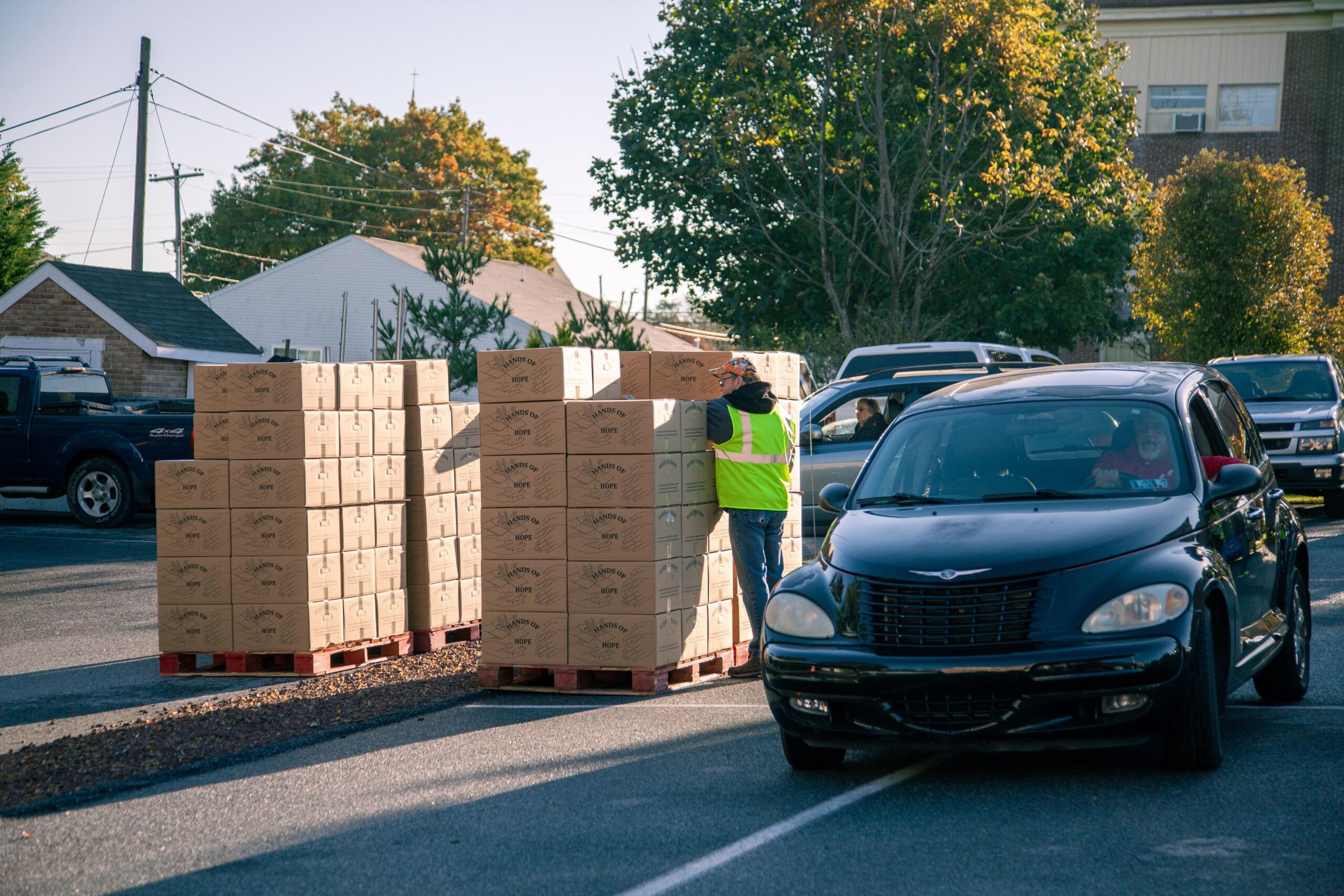 A volunteer in a reflective vest organizes stacked boxes labeled "Hands of Hope" in a parking lot, while cars wait in line for food distribution. Autumn foliage and buildings are visible in the background, indicating a community support event.