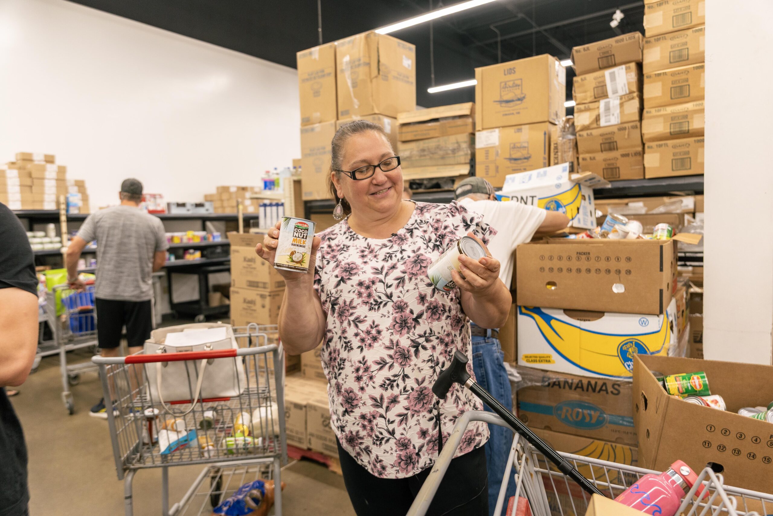 Woman smiling while holding canned goods in a food pantry, surrounded by shelves of food and other shoppers.