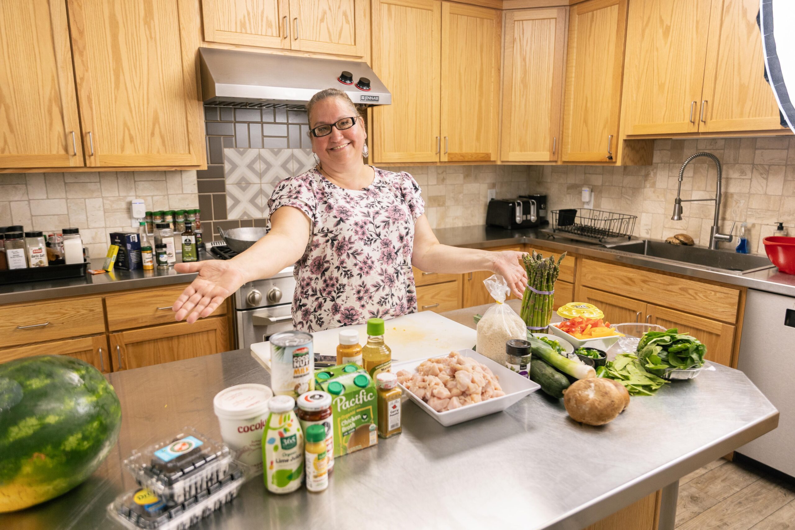 Smiling woman in a kitchen presenting fresh ingredients for cooking, including chicken, vegetables, and spices, on a stainless steel countertop.