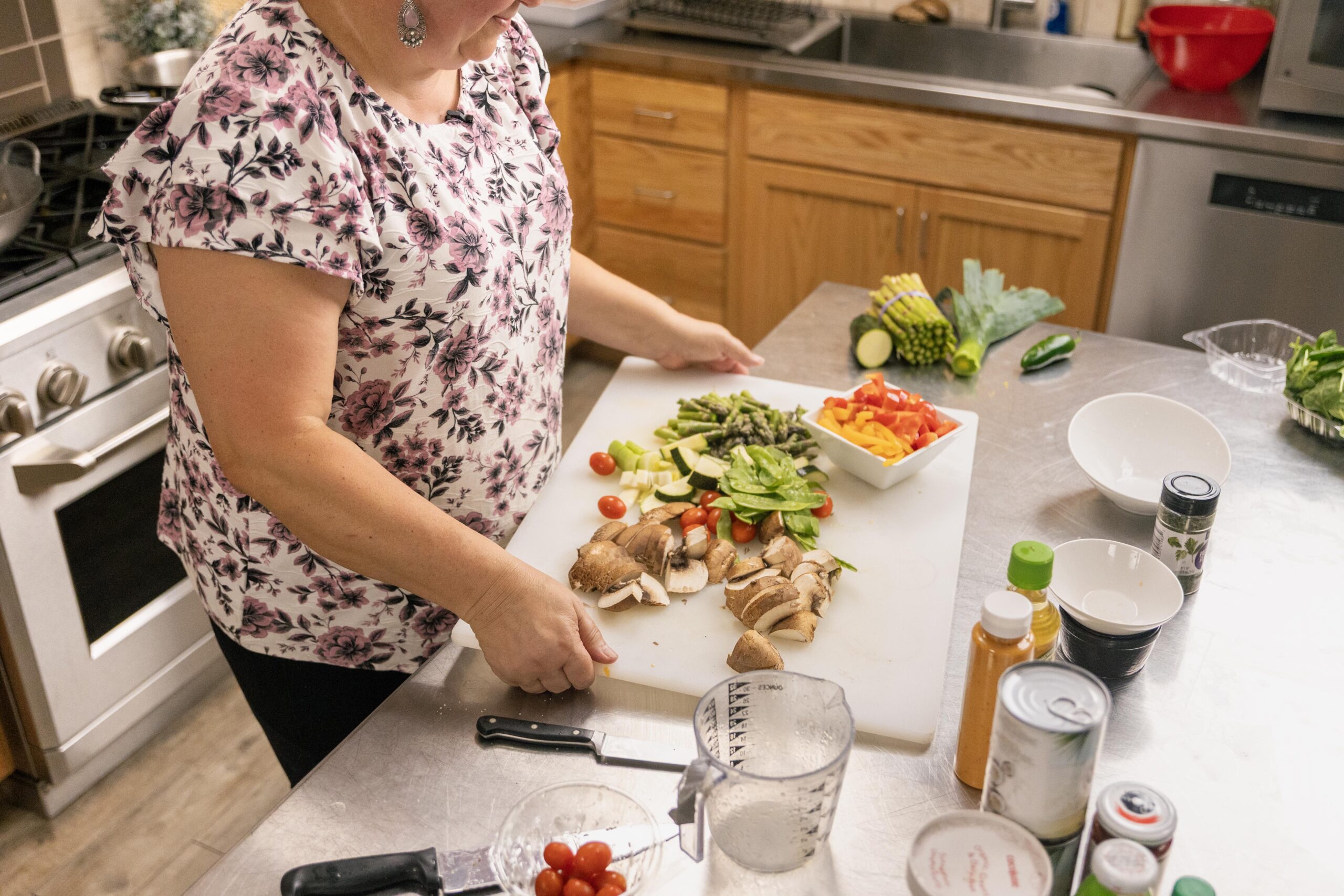 Woman preparing a variety of fresh vegetables, including mushrooms, tomatoes, asparagus, and bell peppers, on a cutting board in a modern kitchen. Cooking utensils and ingredients are visible, highlighting a healthy meal preparation process.